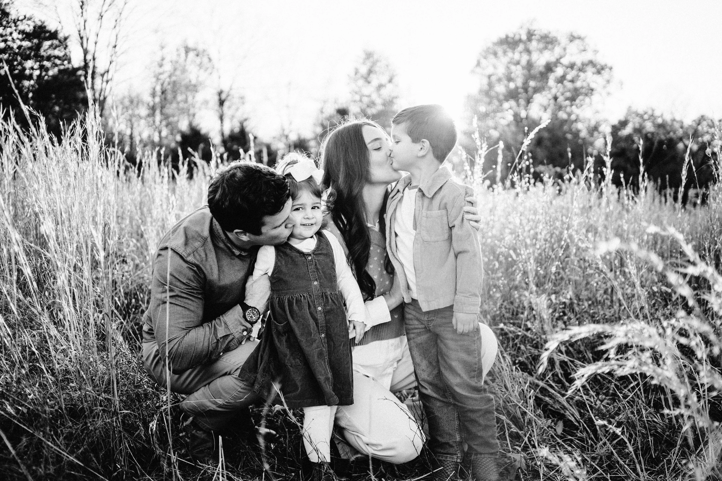 A black and white photo of a family in a grassy outdoor field, sharing a kiss with children smiling.