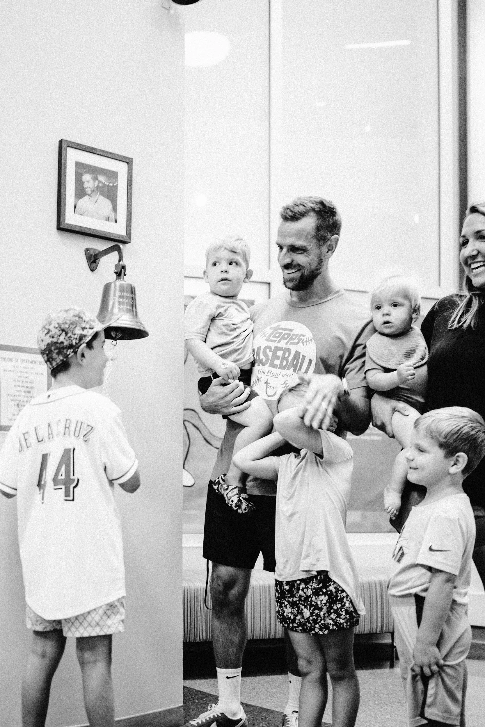 Group of children and adults at an indoor gathering, with one man holding a young boy and surrounded by other children. The scene appears casual, with some children wearing sports jerseys and others in casual clothing.