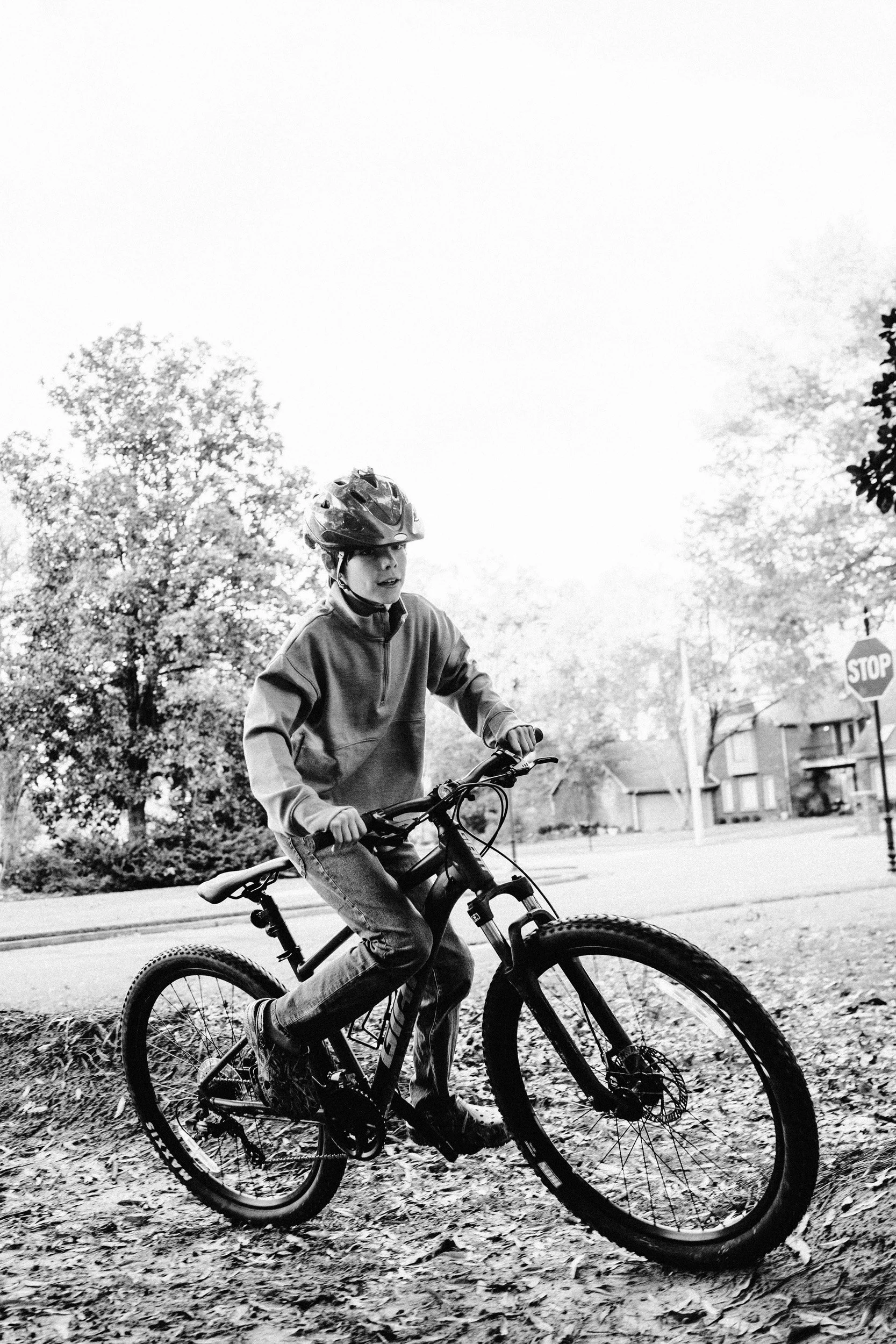 A boy wearing a helmet riding a mountain bike on a trail covered with leaves in a residential neighborhood, with trees and houses in the background.