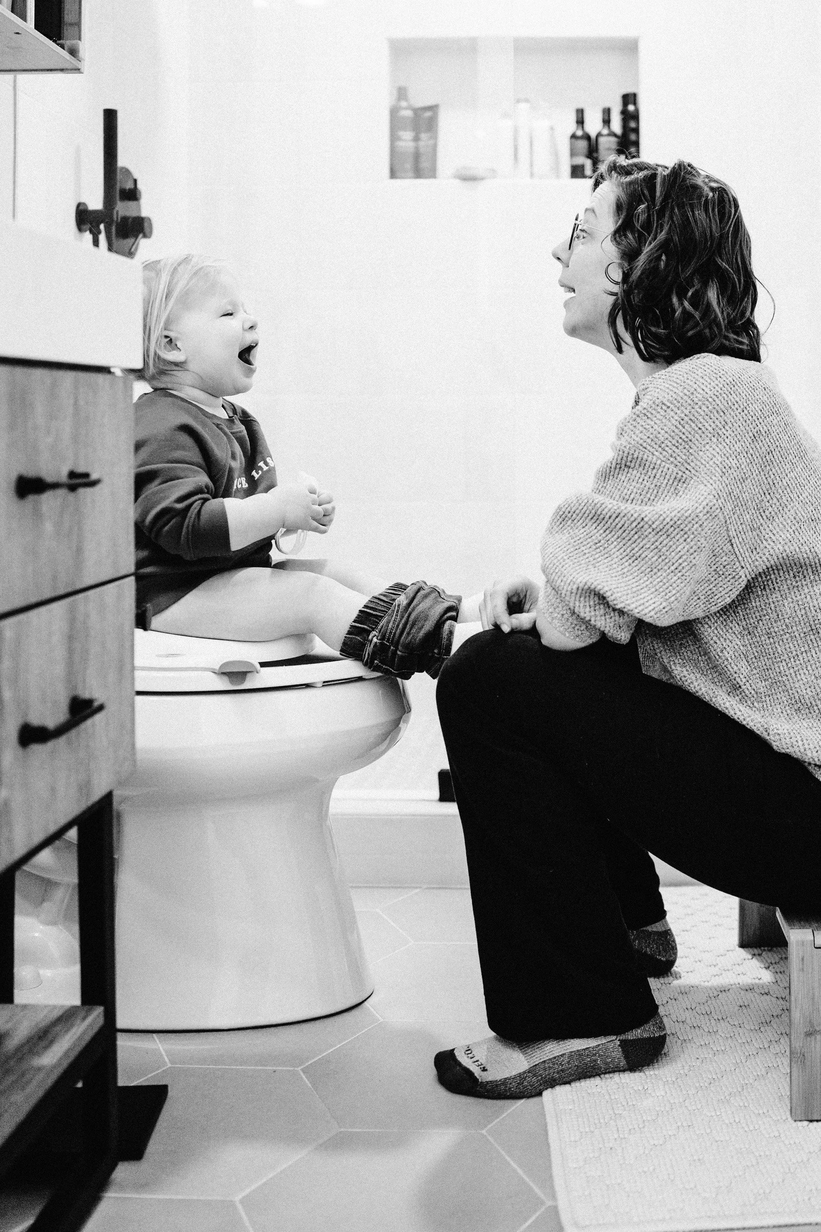 A young child sitting on a potty seat with a big smile, interacting with a woman who is kneeling in front of them, in a bathroom.
