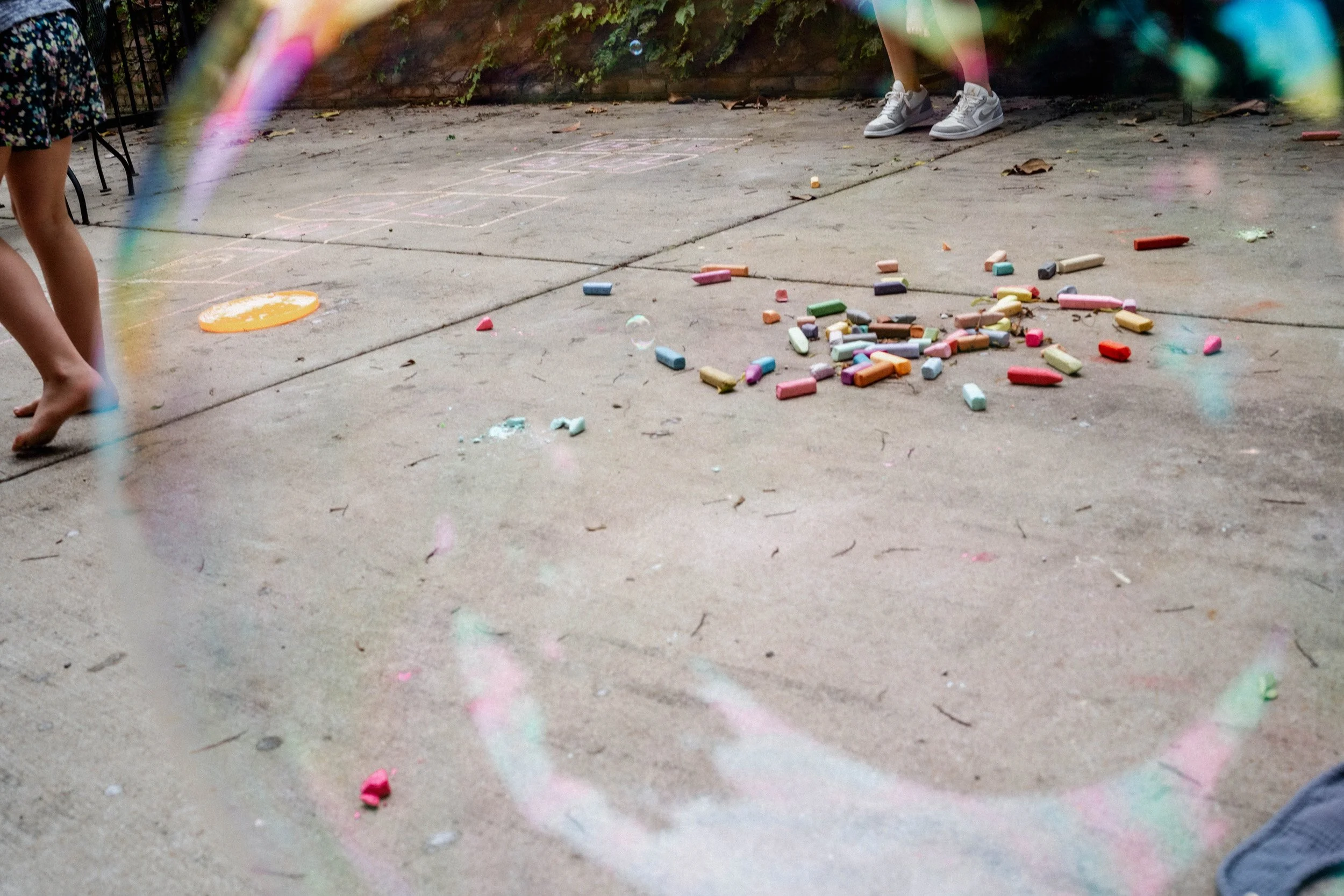 Children playing hopscotch on a concrete driveway, chalk drawings and scattered chalk pieces visible, with one child in sneakers and another barefoot.