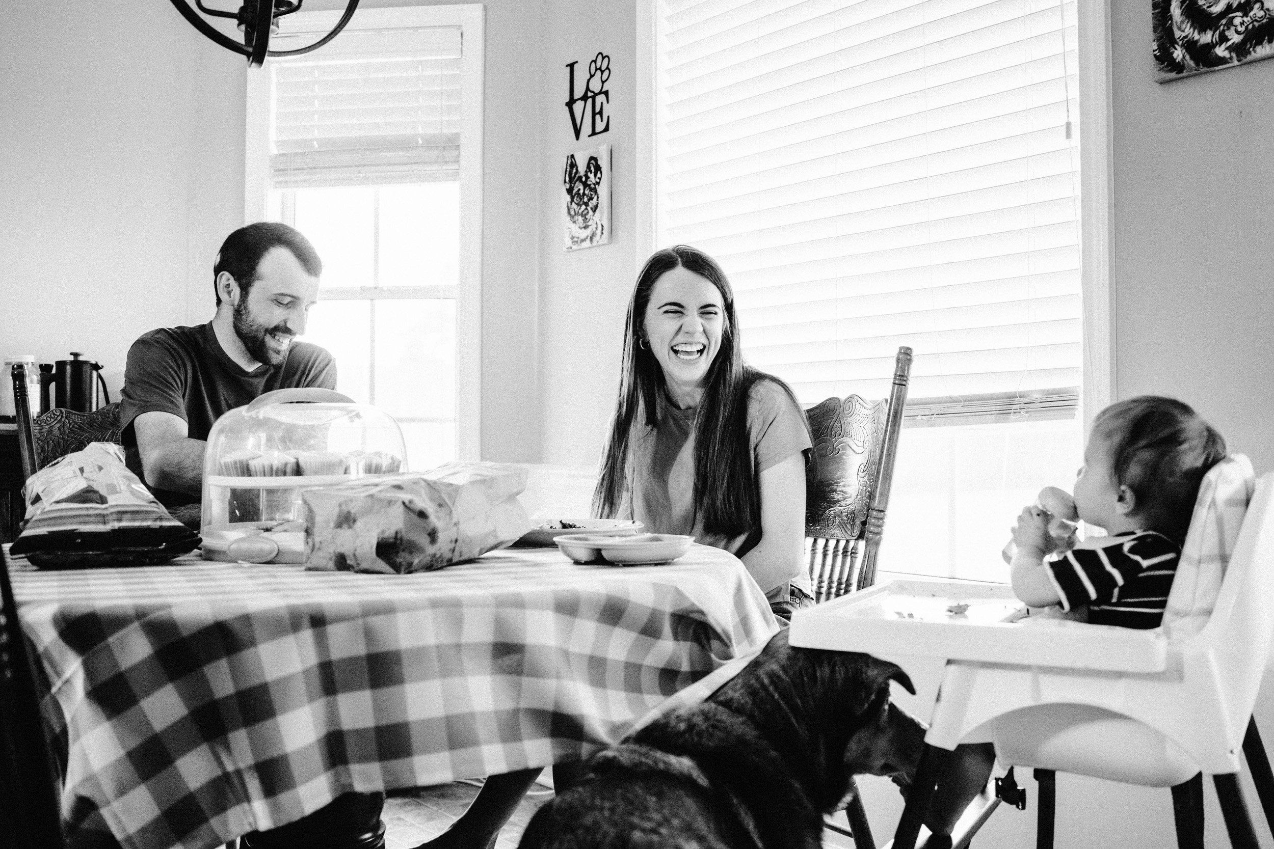 A family enjoying a meal together at a table covered with a checkered tablecloth. The scene includes a man, a woman, and a young boy sitting in a high chair, all smiling and laughing. The table has food, dishes, and a cake. Light streams in through large windows with blinds in the background, and wall decor, including a