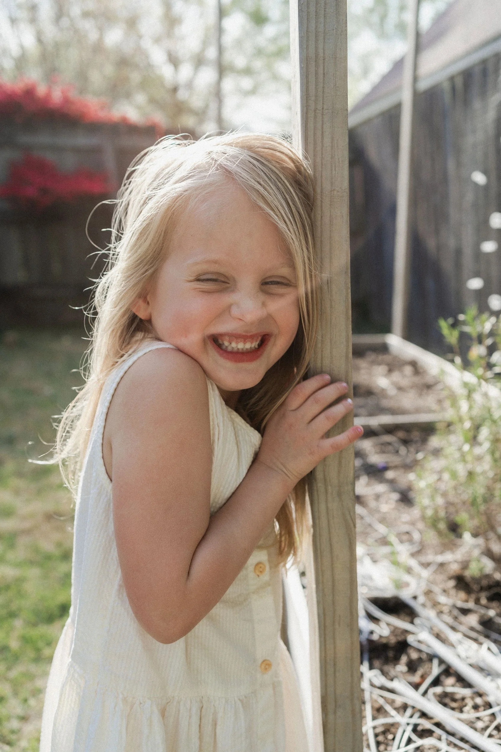 A young girl with long blonde hair and a big smile, standing outdoors next to a wooden post, wearing a white sleeveless dress, in a backyard with blurred trees and a fence in the background.