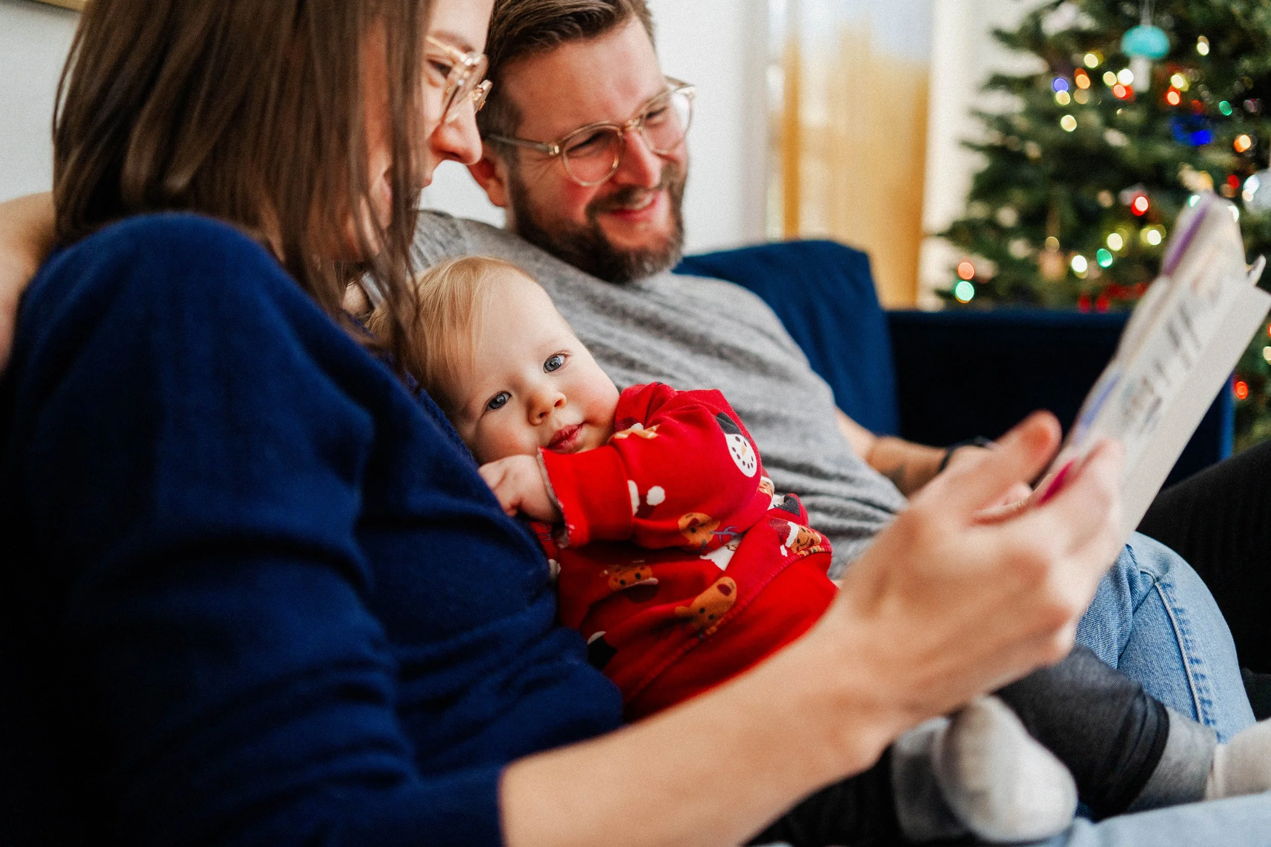 A family sitting on a couch during Christmas, looking at a tablet. An adult and a child are close, and a baby is resting on the adult's lap. A decorated Christmas tree is visible in the background.