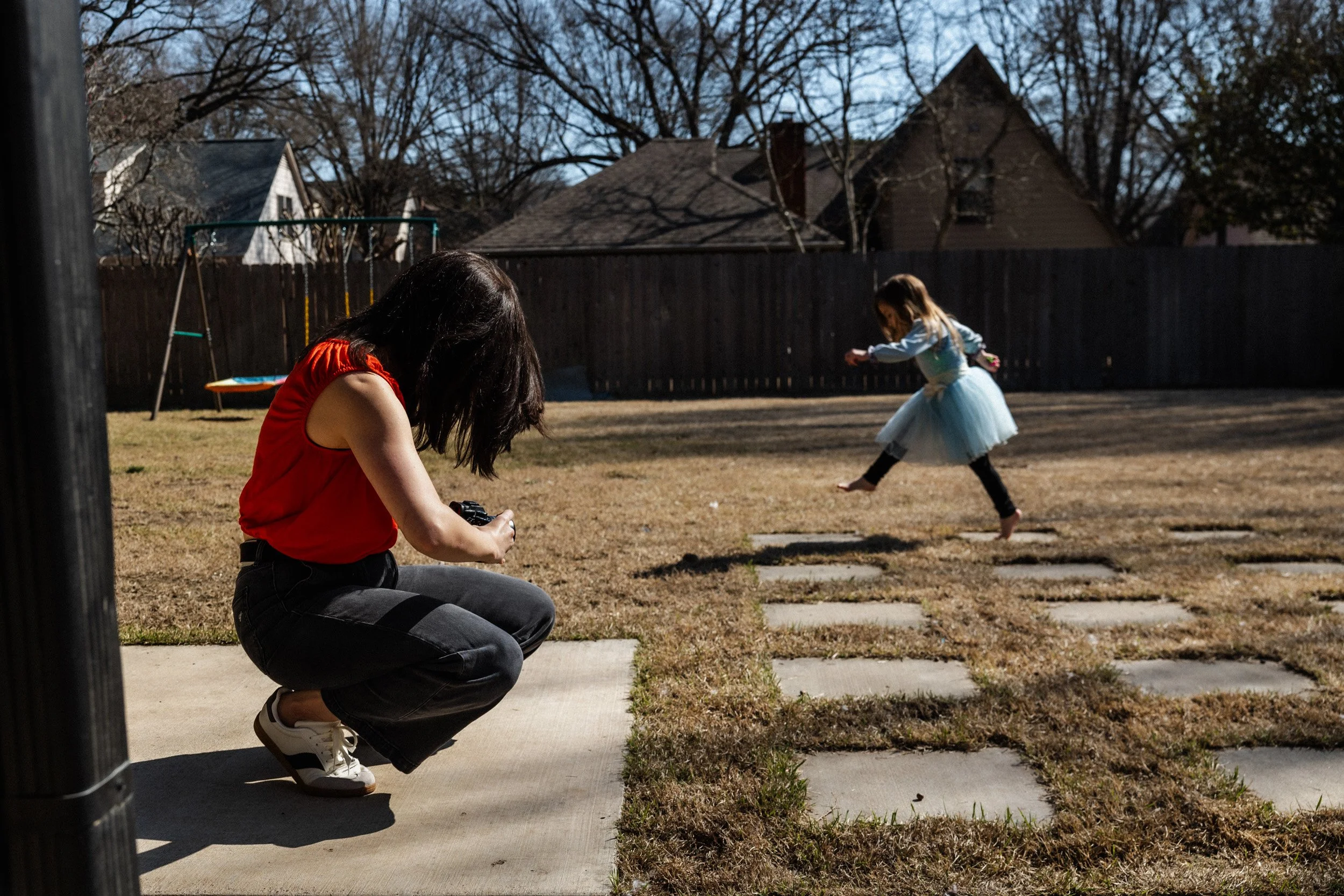 A woman crouching on a concrete sidewalk taking a photograph of a girl in a blue dress who is running in a backyard with a wooden fence, trees, and houses in the background.