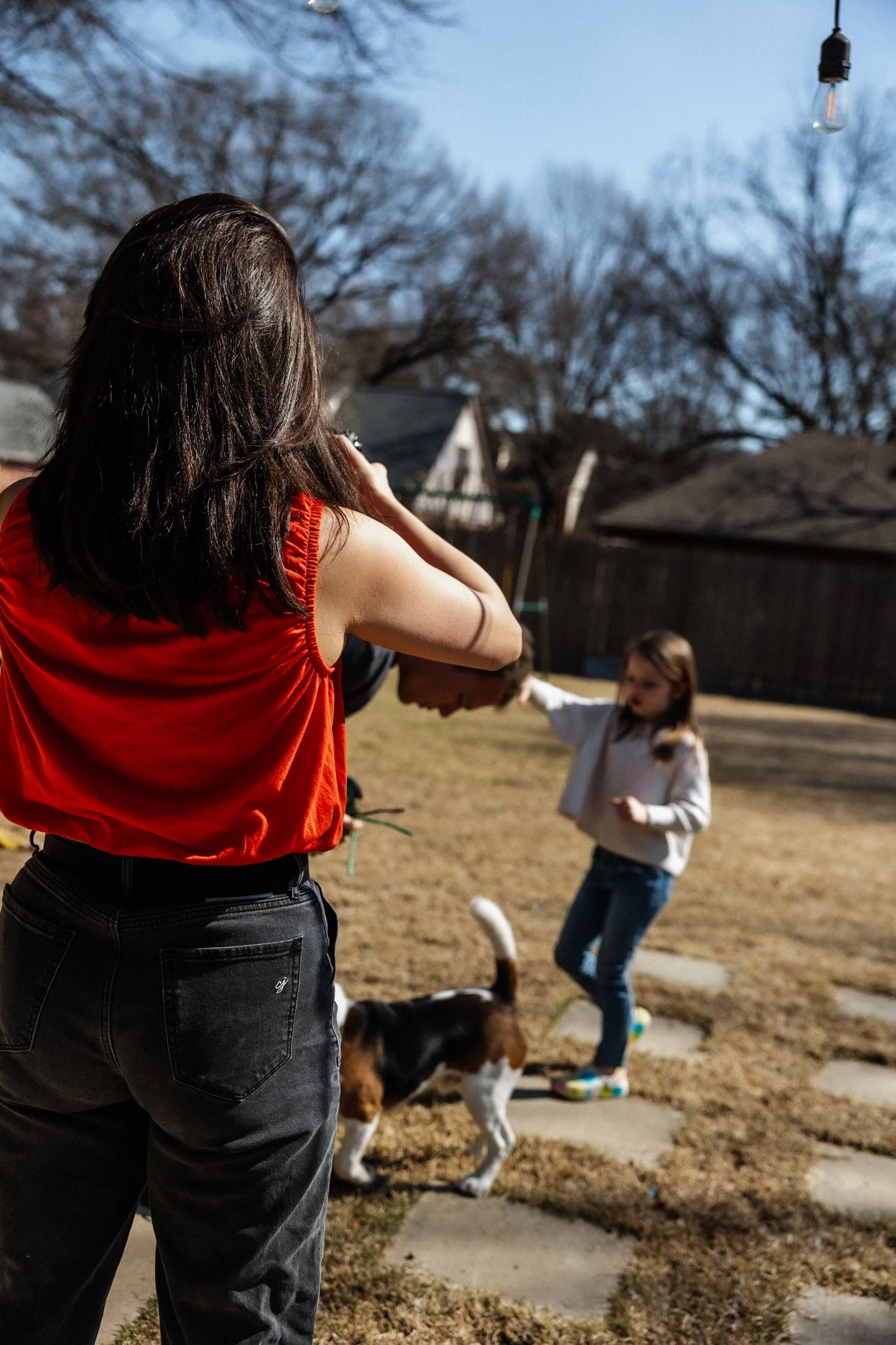 A woman with long dark hair wearing a red sleeveless top and black jeans is outdoors, holding a camera or phone, taking a picture of children and dogs playing in a yard. The yard has dry grass, a sidewalk, and some trees in the background. The children include a girl with glasses and a white hoodie.