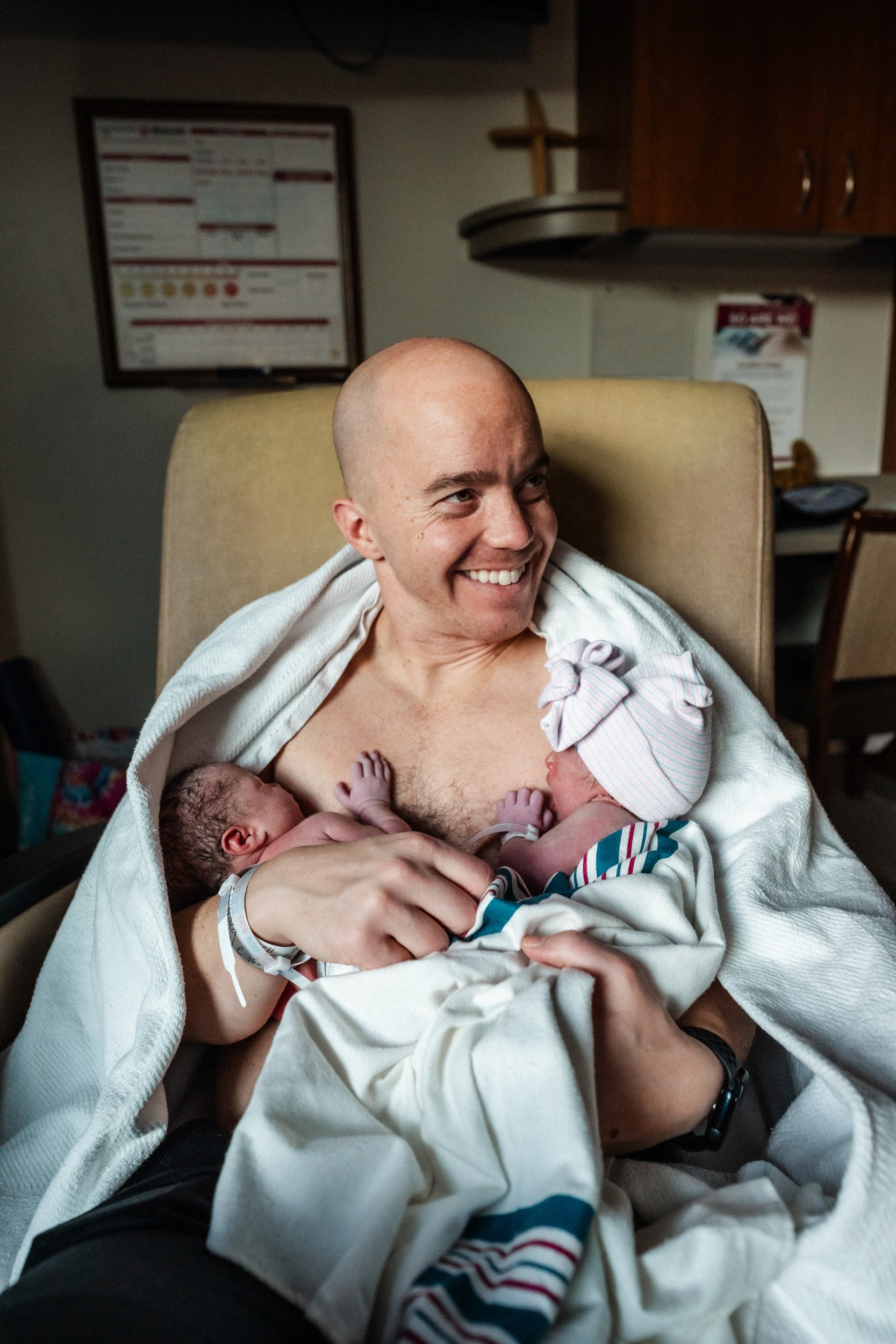 A smiling man holding two newborn babies in a hospital room.