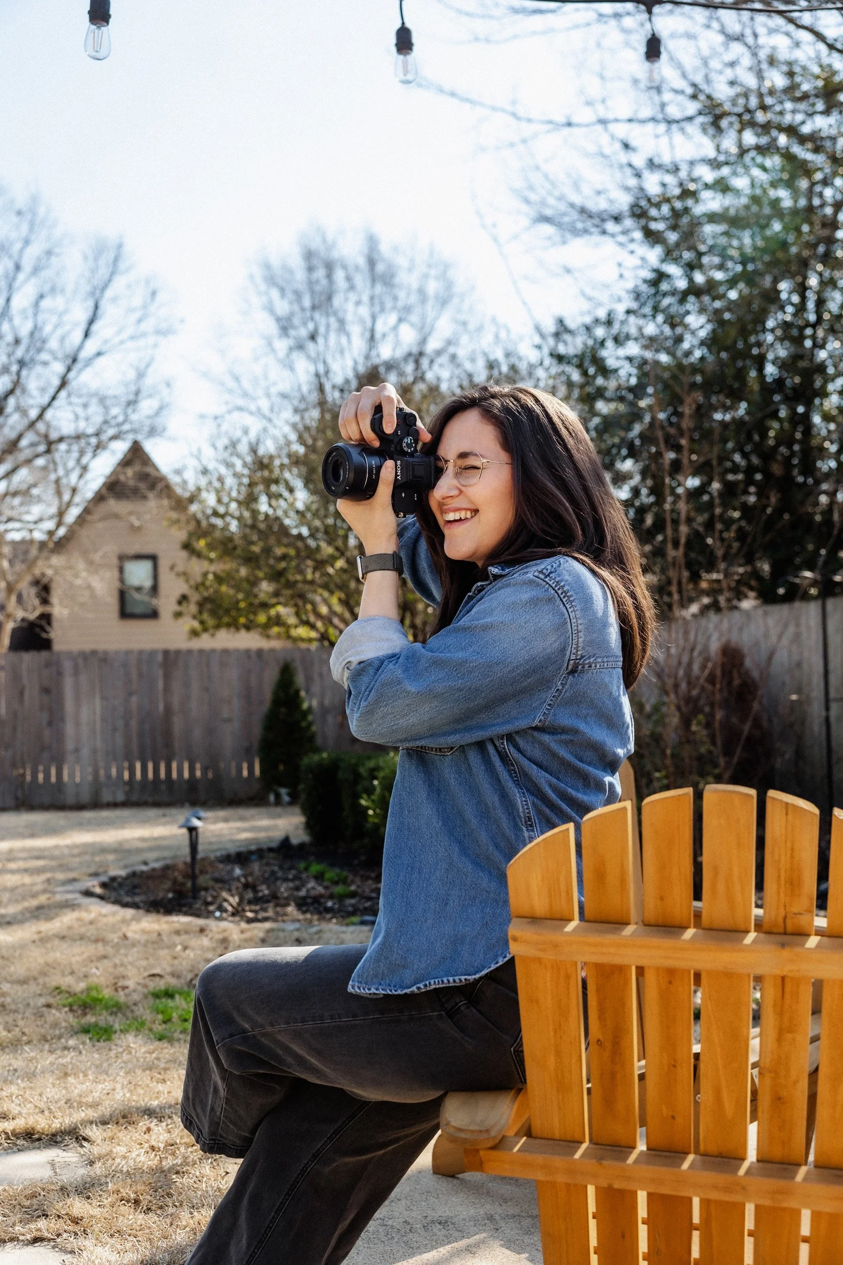 A woman with dark hair, glasses, and a denim jacket, sitting on a wooden chair outdoors, taking a photograph with a camera, with a background of trees, a house, and hanging string lights.