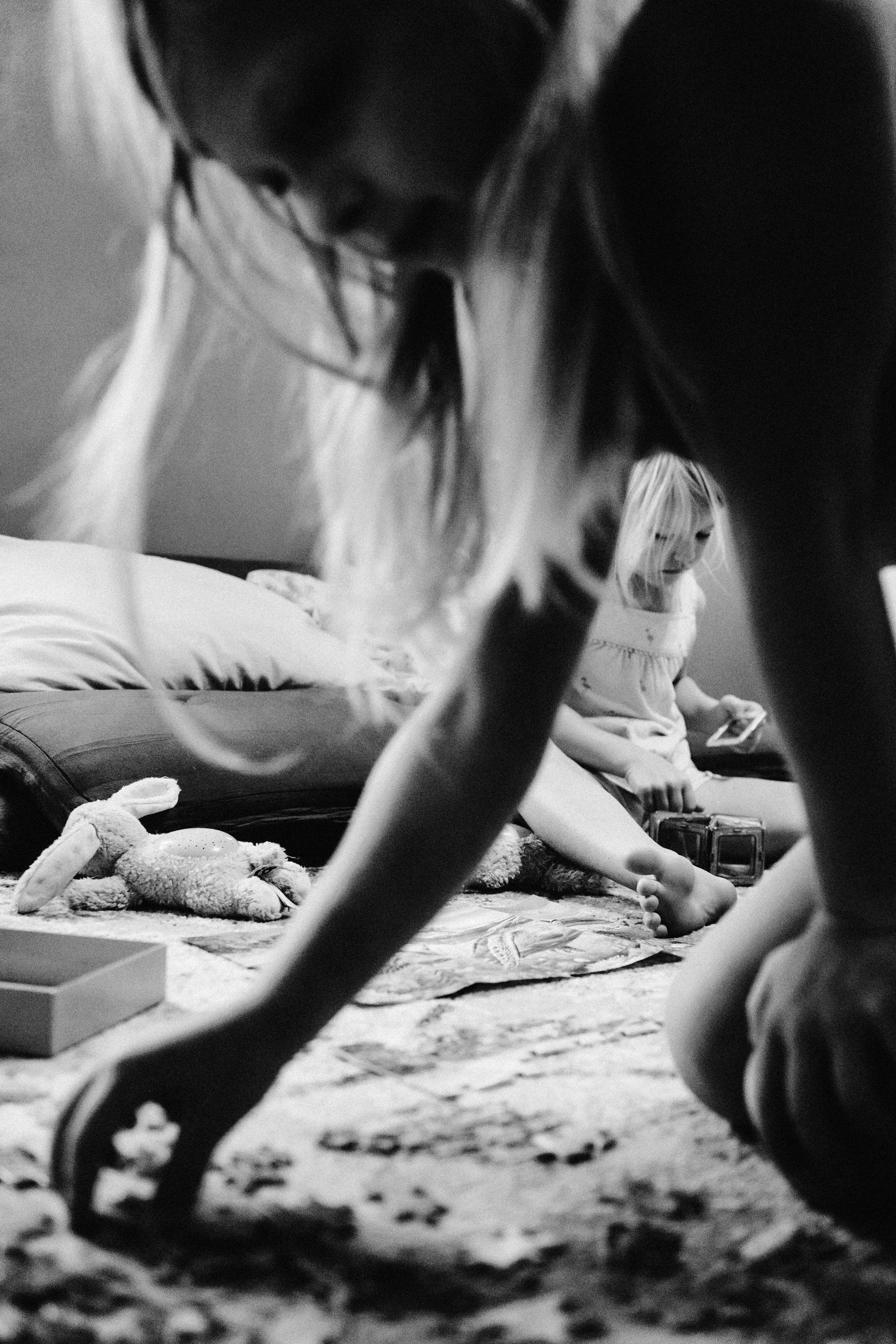A child sitting on the floor and playing with a toy truck, with another child lying in the background using a mobile device, in a room with a bed and stuffed animals.