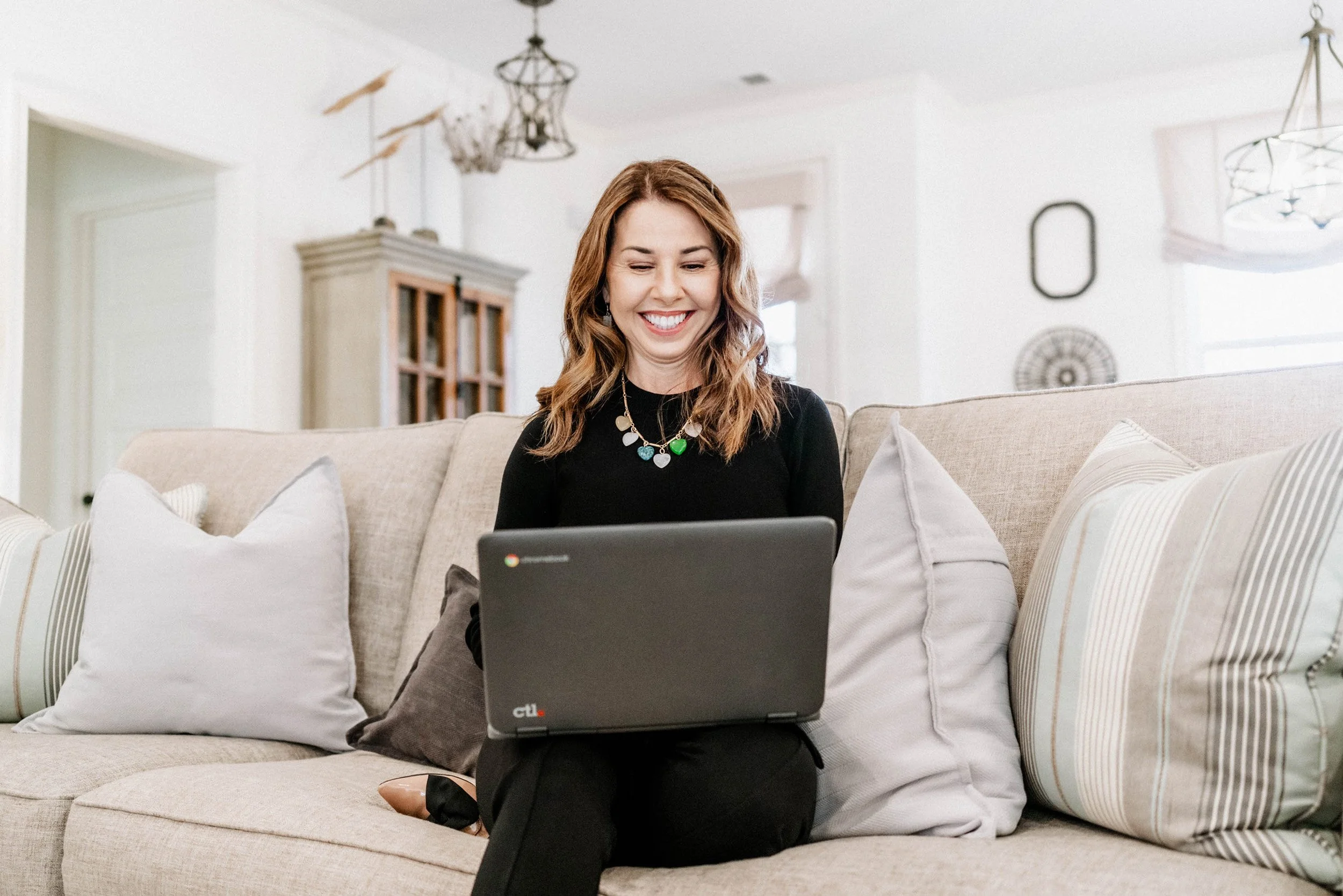 A woman with wavy brown hair smiling while sitting on a beige sofa, using a laptop, in a bright living room with decorative ceiling lights and a wooden cabinet in the background.