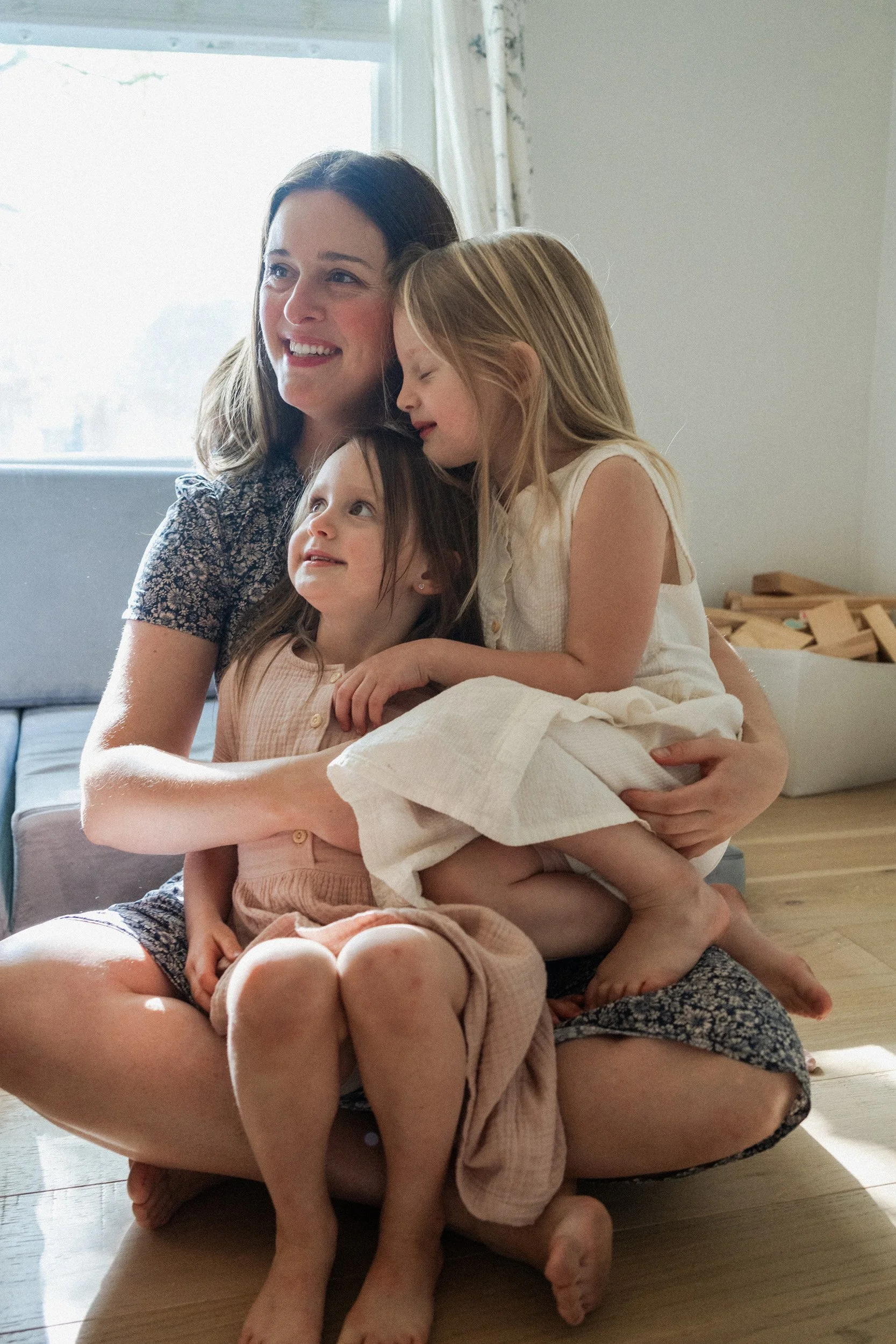 A woman and three young girls sitting on the floor near a large window, embracing and smiling together in a cozy, well-lit room.