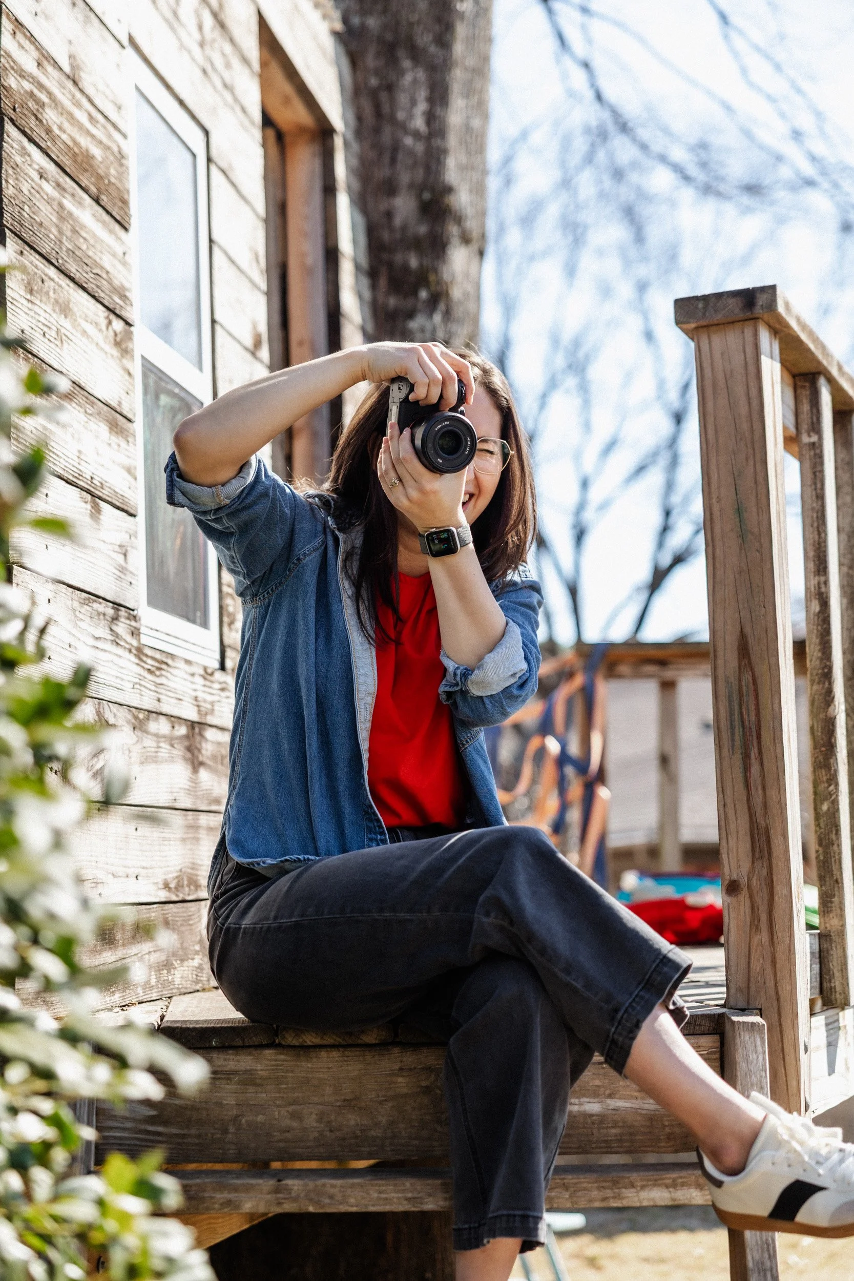 Young woman sitting on wooden steps outside, holding a camera and taking a photo, wearing glasses, a denim jacket, red shirt, black pants, and white sneakers, with a tree and house in the background on a sunny day.