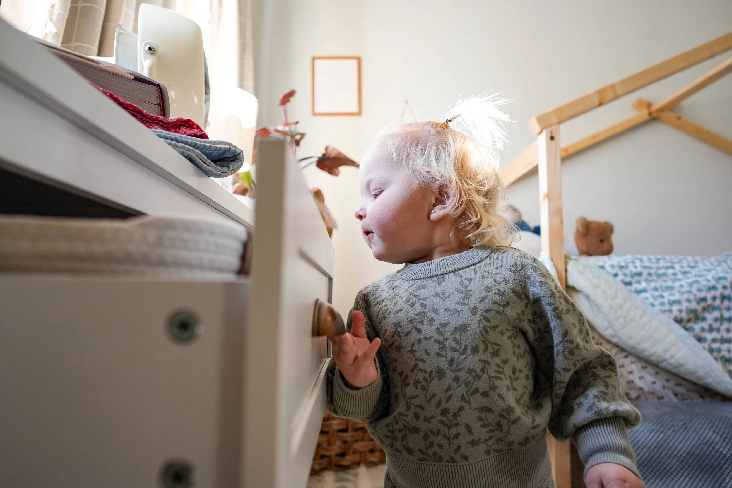 A young child with blonde hair, wearing a patterned gray sweater, is standing beside a white dresser, looking at objects on top, in a brightly lit bedroom.
