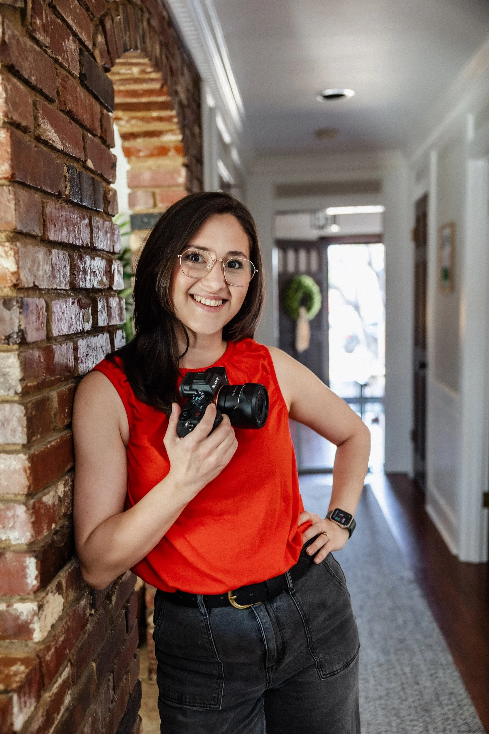 A woman with brown hair, glasses, and a bright smile standing indoors near a brick wall, wearing a red sleeveless top, black jeans, and a smartwatch, holding a camera in her right hand.