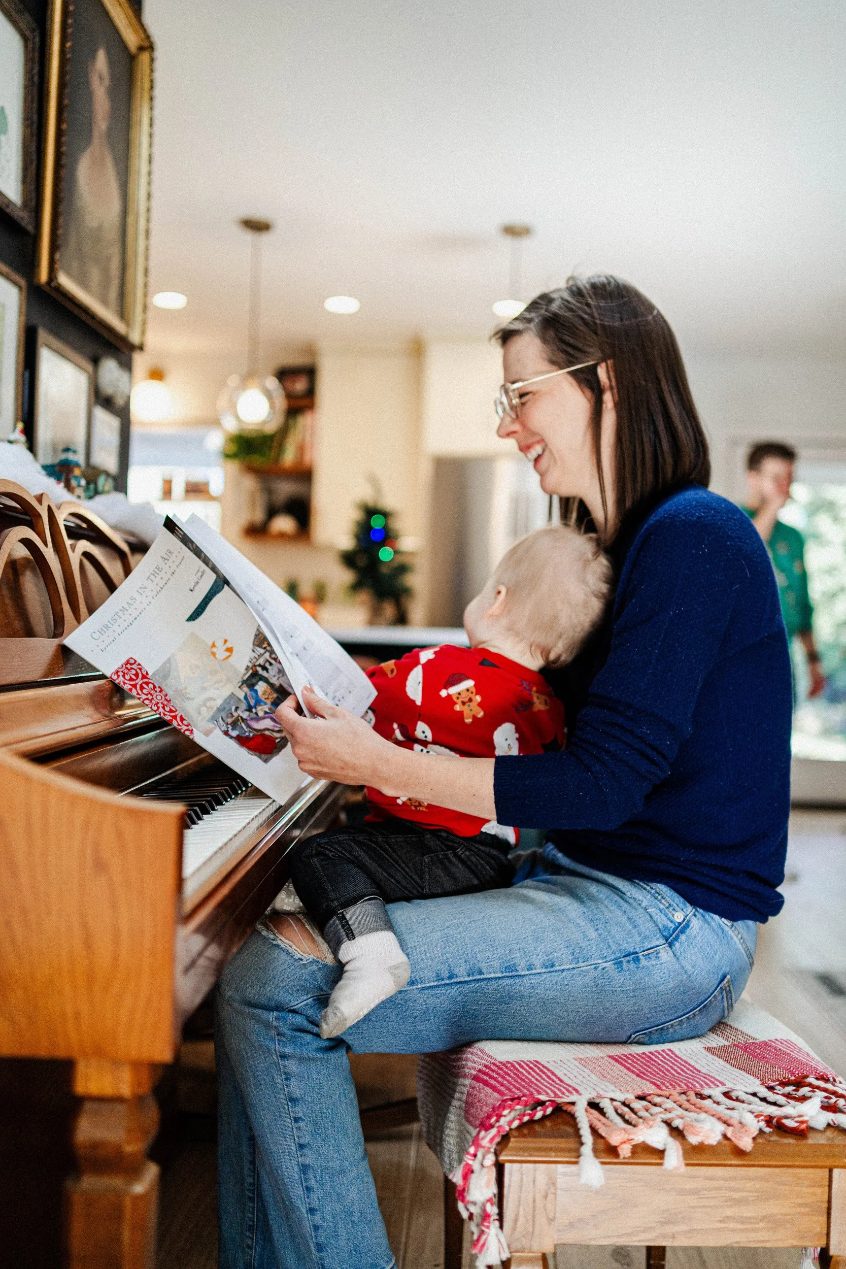 A woman with glasses and wearing a blue sweater is sitting on the floor playing the piano while a young child in Christmas pajamas sits on her lap and looks at the music on the piano.