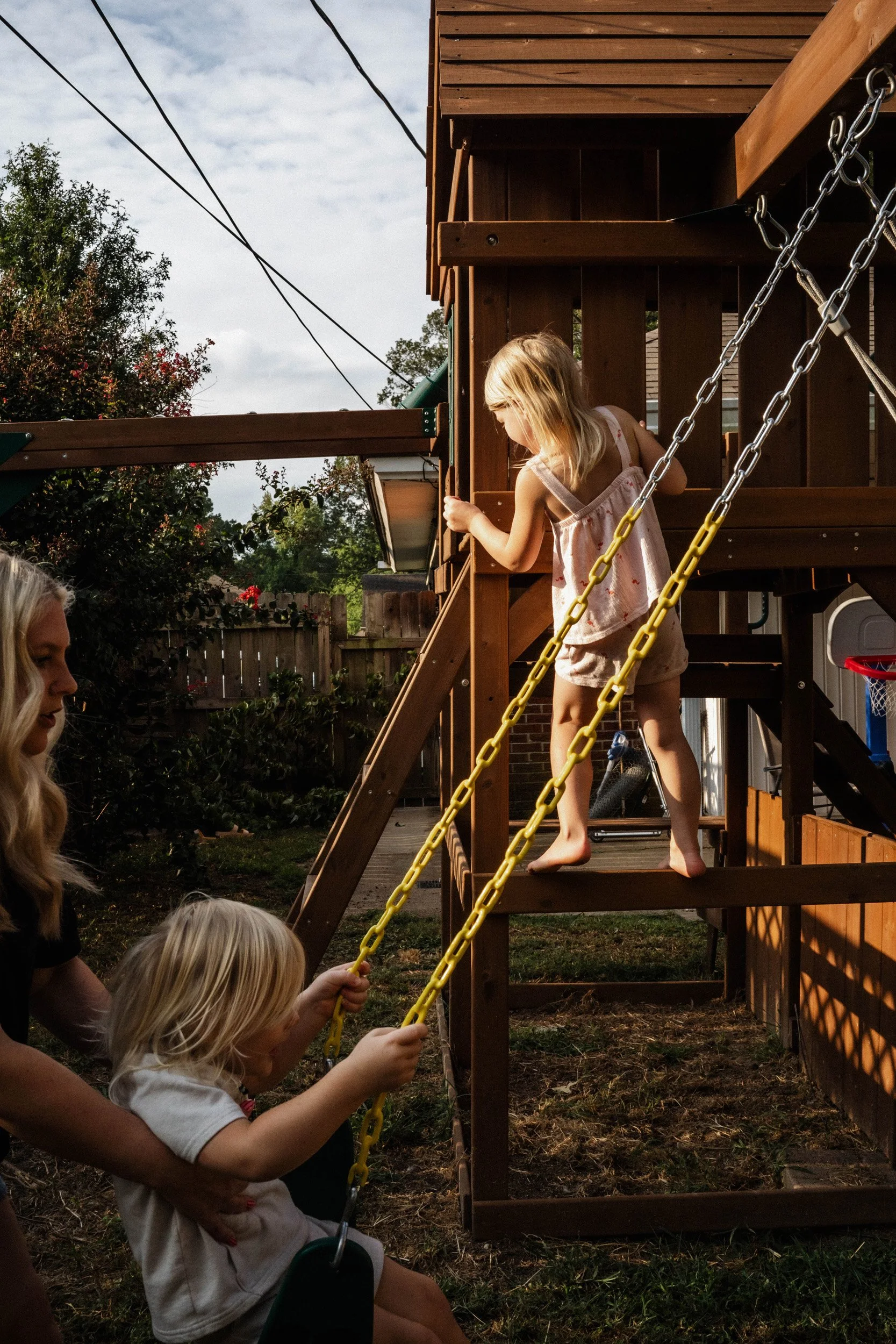 A group of children playing on a wooden outdoor playset in a backyard during daytime. A girl with blonde hair in a pink dress is climbing on the playset, while others are holding the yellow chains of the swing.