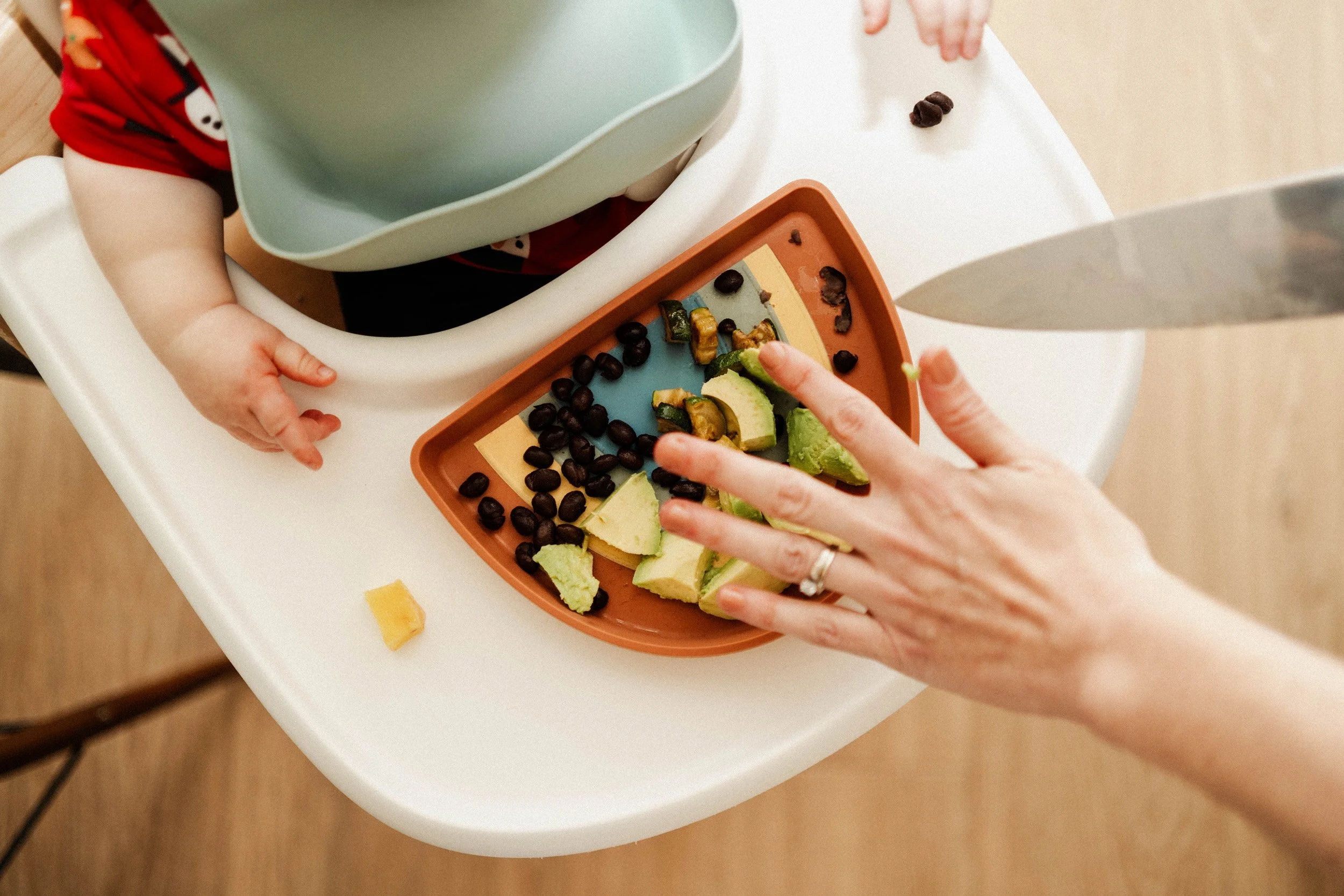 Child and adult preparing a snack with sliced avocado, black beans, and other vegetables or fruits on a table.