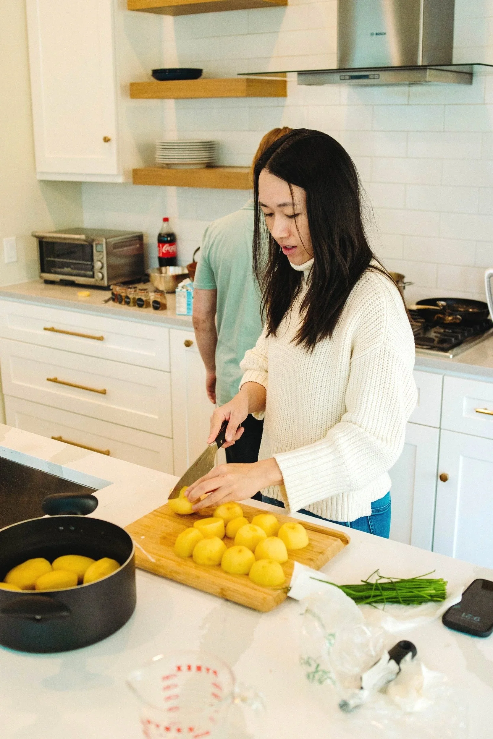 A woman with long dark hair, wearing a cream-colored sweater, is slicing peeled potatoes on a wooden cutting board in a modern kitchen. Other peeled potatoes are on the cutting board. In the background, a man with light brown hair, wearing a green shirt, is facing away. The kitchen counter holds a black pot, a bottle of Coca-Cola, and a toaster oven. There are white cabinets with gold handles and open wooden shelves with dishes. On the white countertop, there are some green herbs, crumpled paper towels, and a phone.