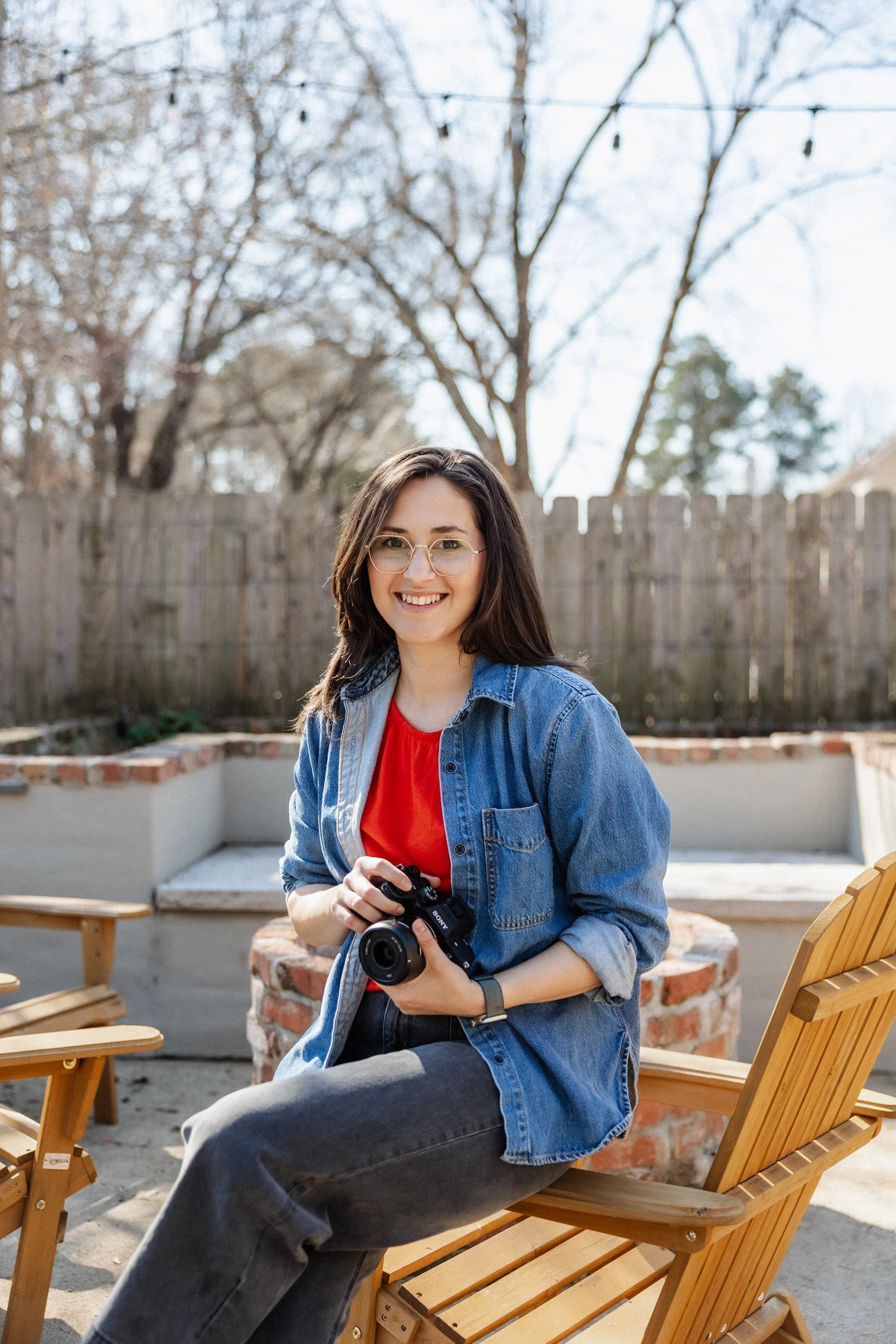 A young woman with glasses sitting on a wooden bench outdoors, smiling, holding a camera, with leafless trees and a wooden fence in the background.