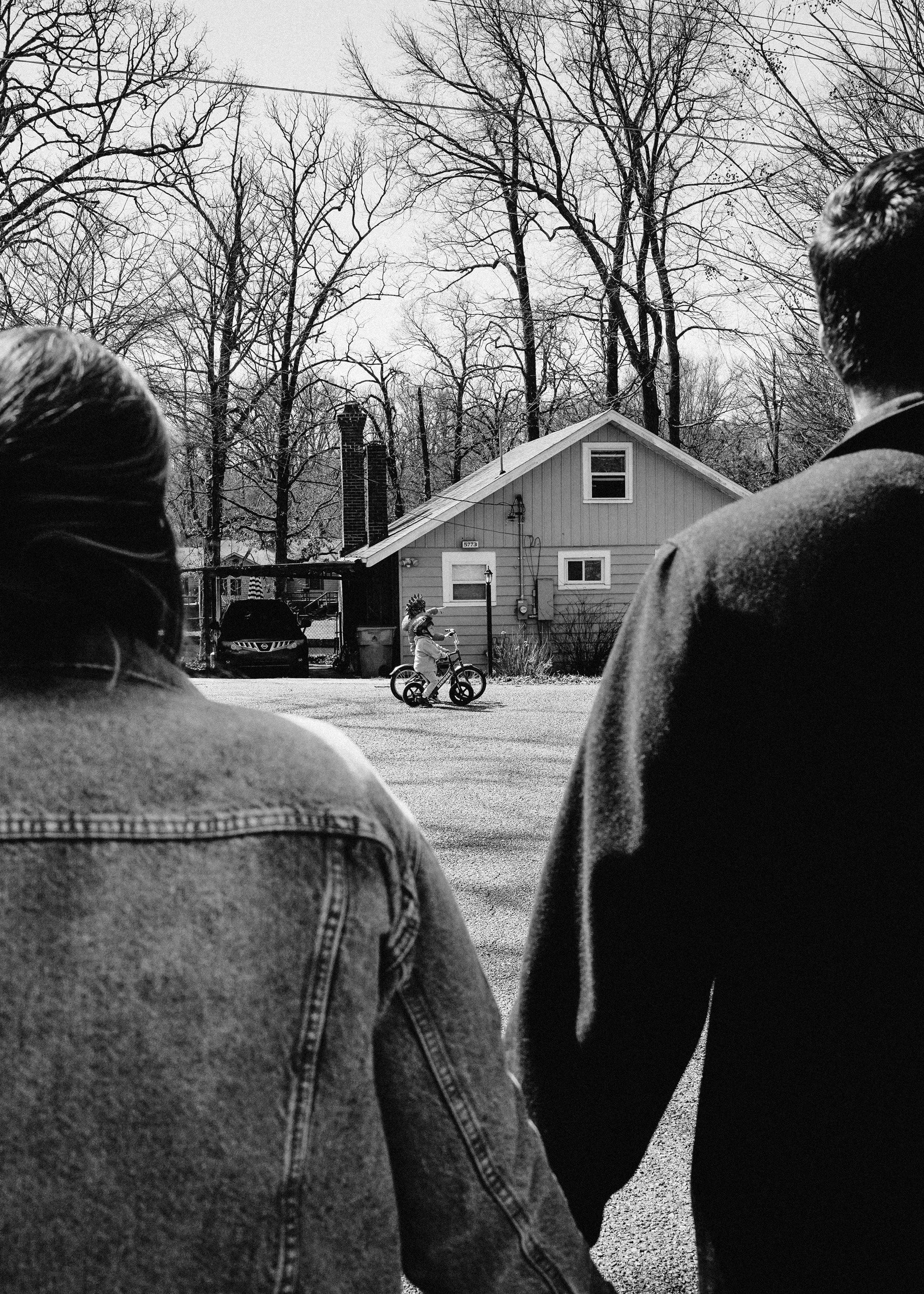 People standing outdoors watching a girl riding a bicycle on a neighborhood street with houses and leafless trees in the background