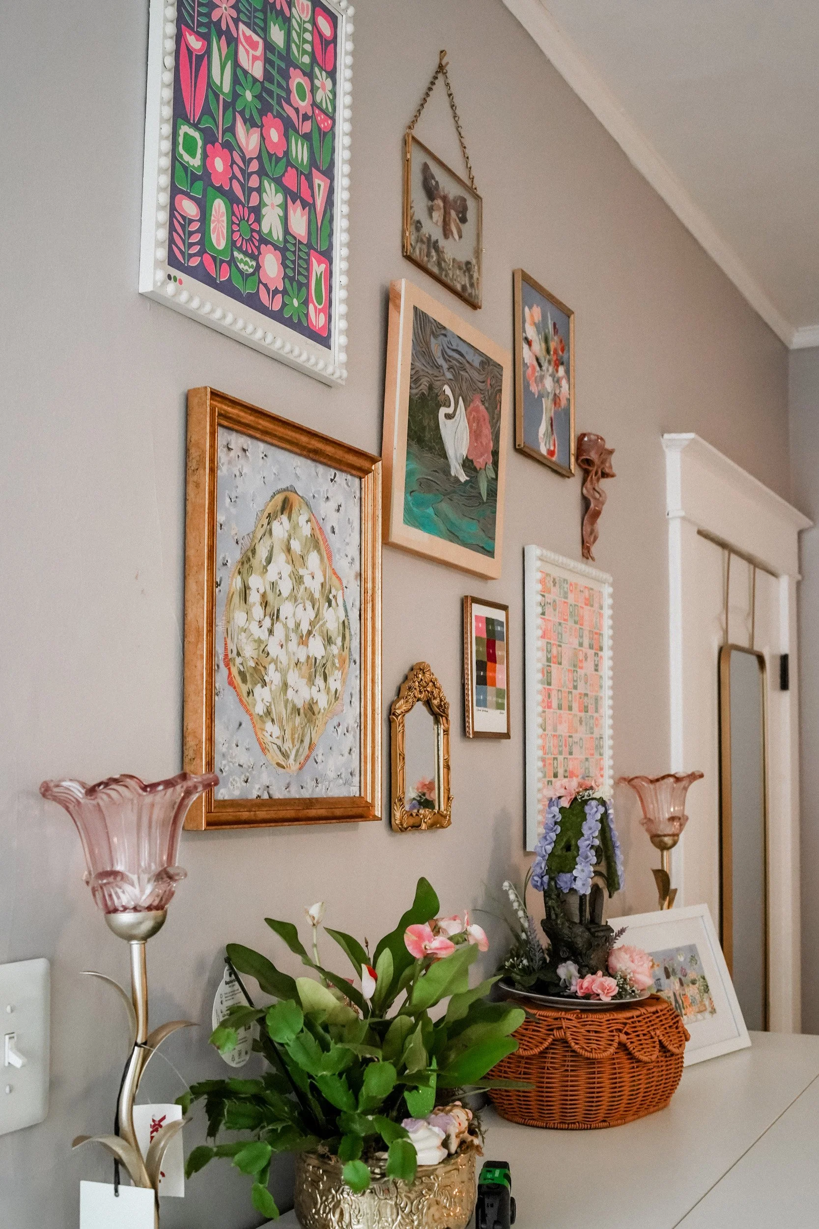 A wall decorated with various framed art pieces, including floral prints, a butterfly, and a white swan, with pink glass flower lamps on a table below and a basket of flowers nearby.
