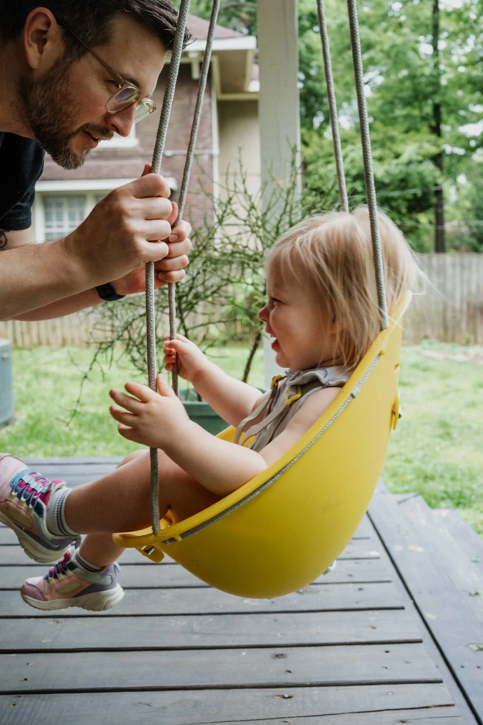 A man with glasses and a beard gently holding hands with a young girl sitting on a yellow swing outdoors.