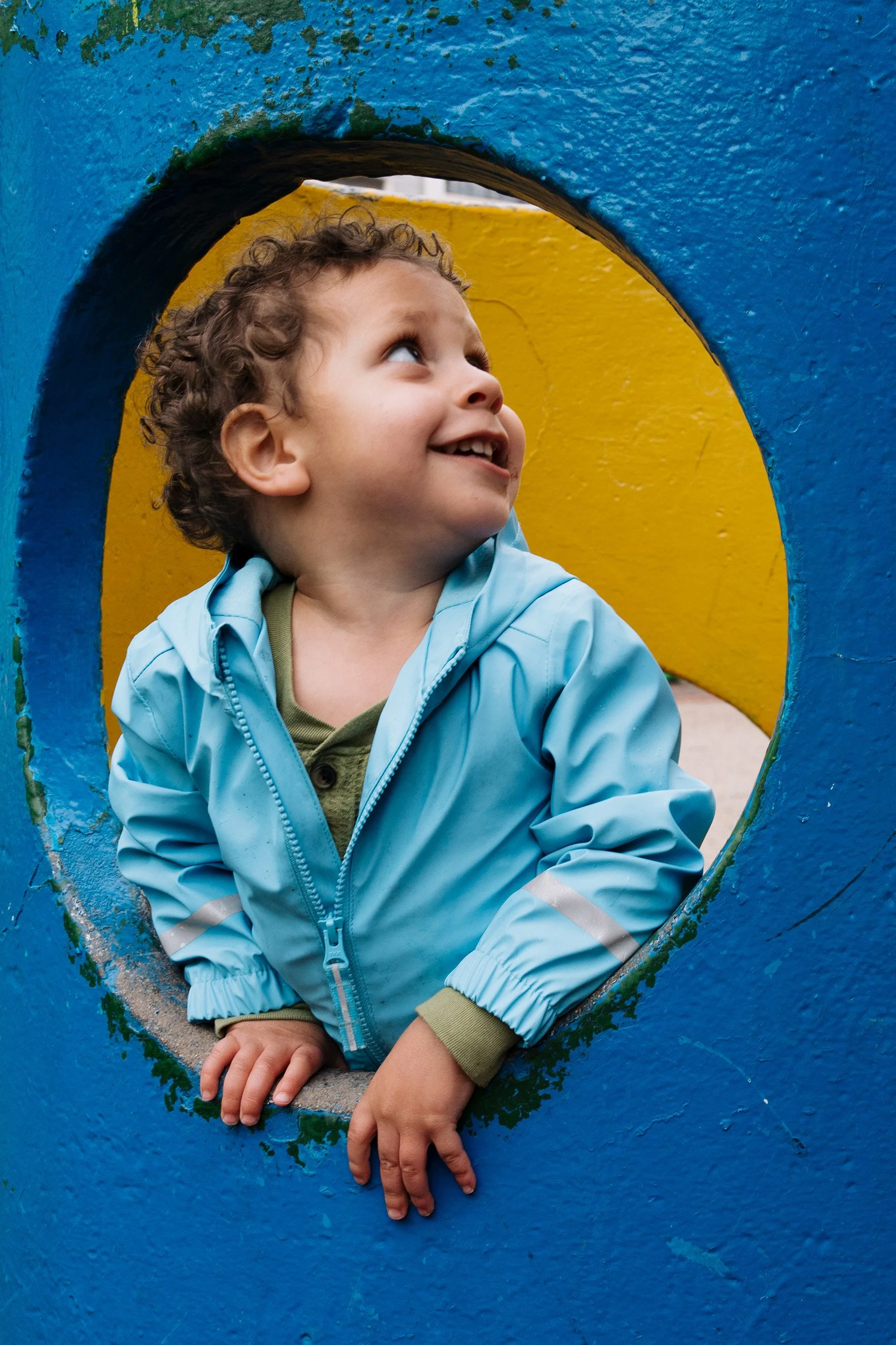 A young boy with curly hair, wearing a light blue jacket, is looking up and smiling through a circular opening in a blue and yellow painted wall or structure.