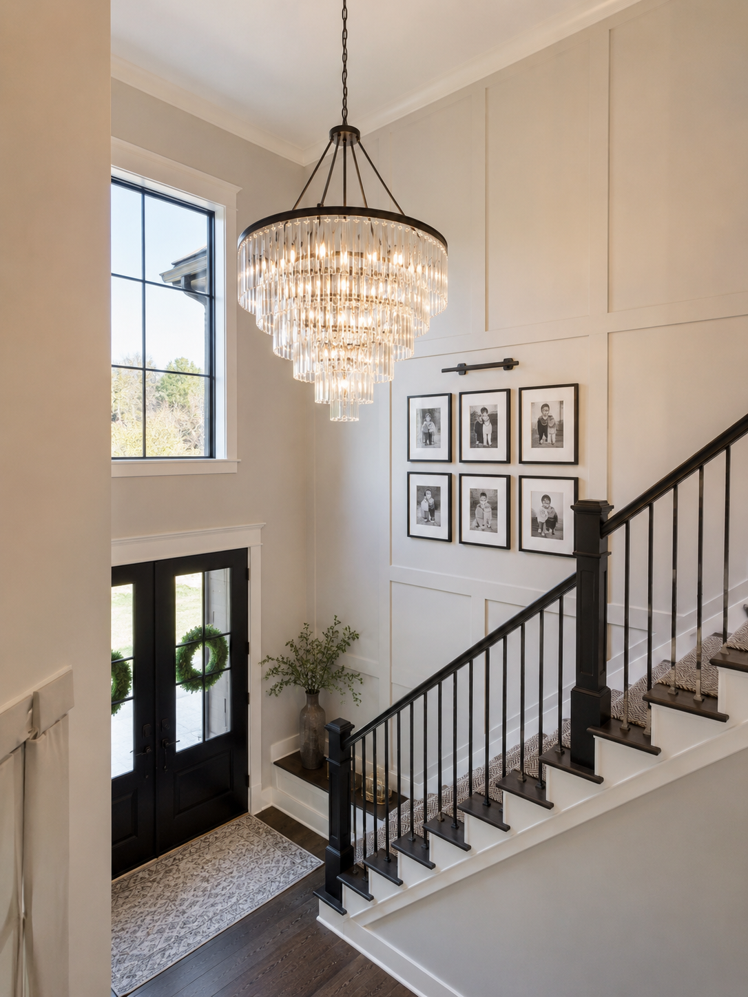 Entryway of a modern home with black front door decorated with wreaths, a staircase with black railing, a large crystal chandelier, black and white framed photos on the wall, a potted plant, and a window letting in natural light.