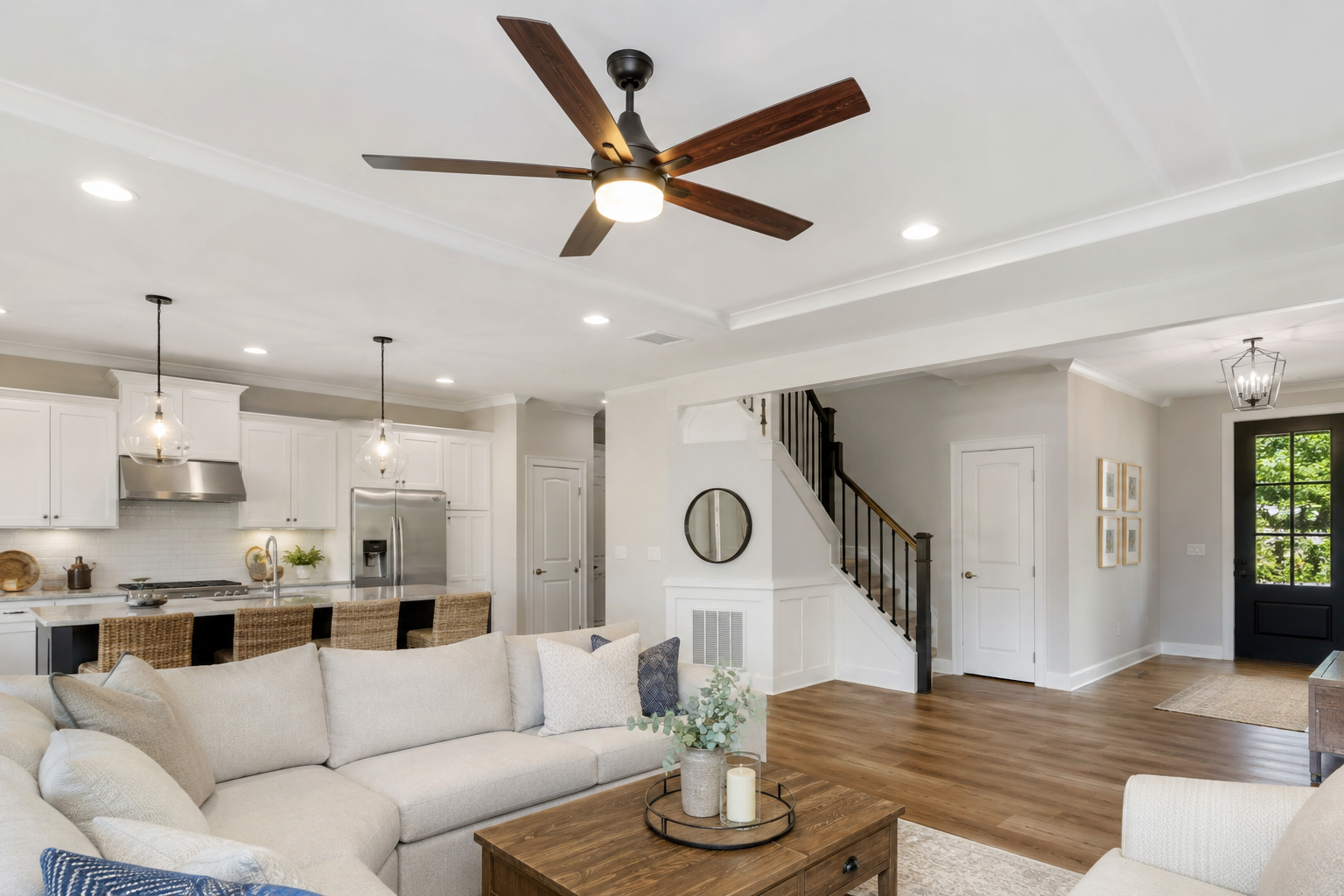 Open-concept living room and kitchen with white cabinets, stainless steel appliances, black staircase, beige couch, wooden coffee table, and hardwood flooring, lit by ceiling lights and a ceiling fan.