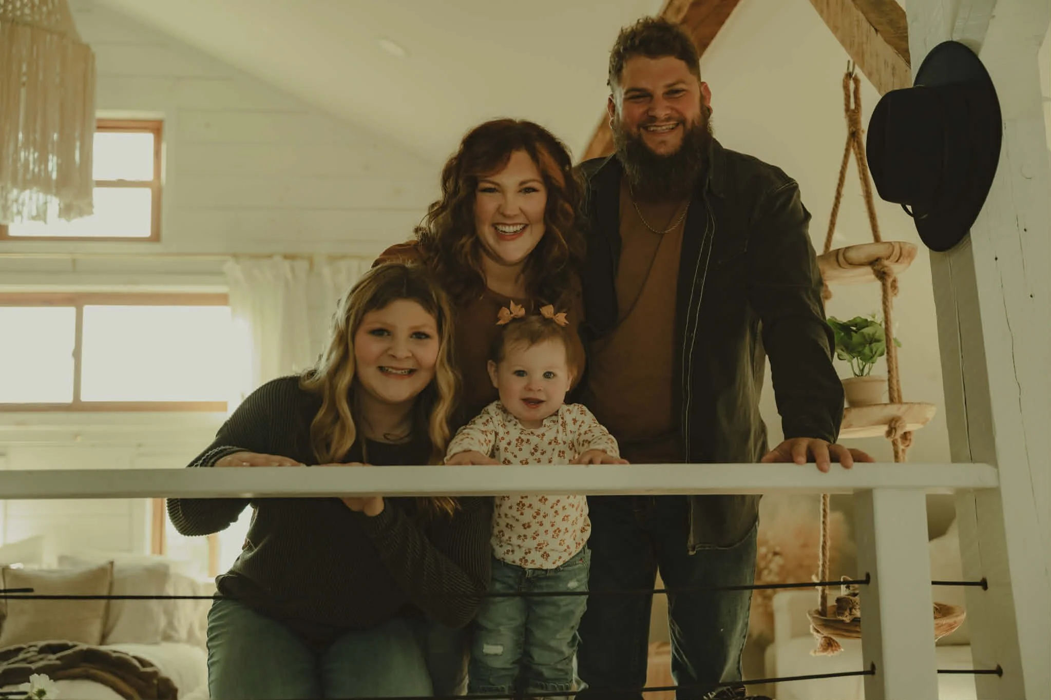 A family of five posing indoors, smiling, with two children in the foreground and two adults behind them, with a wooden staircase and window in the background.