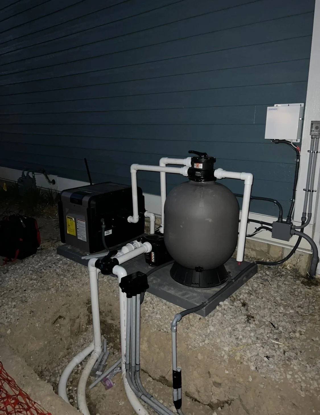 Swimming pool filter system with a large gray tank, white pipes, a pump, and an electrical control box outside against a blue house wall.