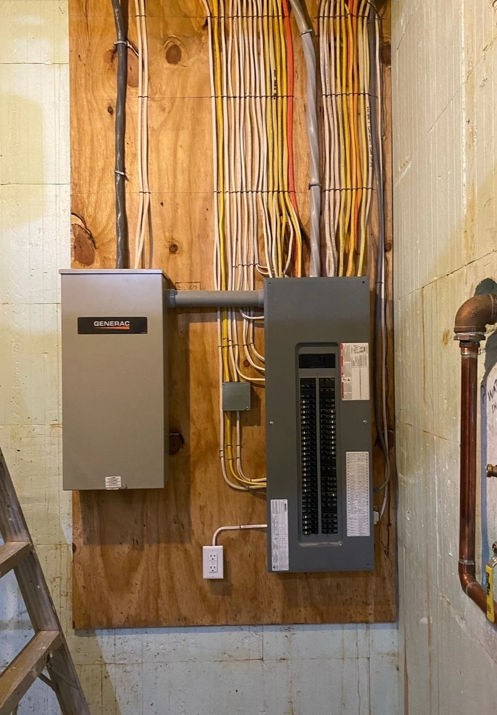 An electrical panel and a generator box installed on a wooden board with multiple electrical wires connected, in what appears to be a basement or utility room.