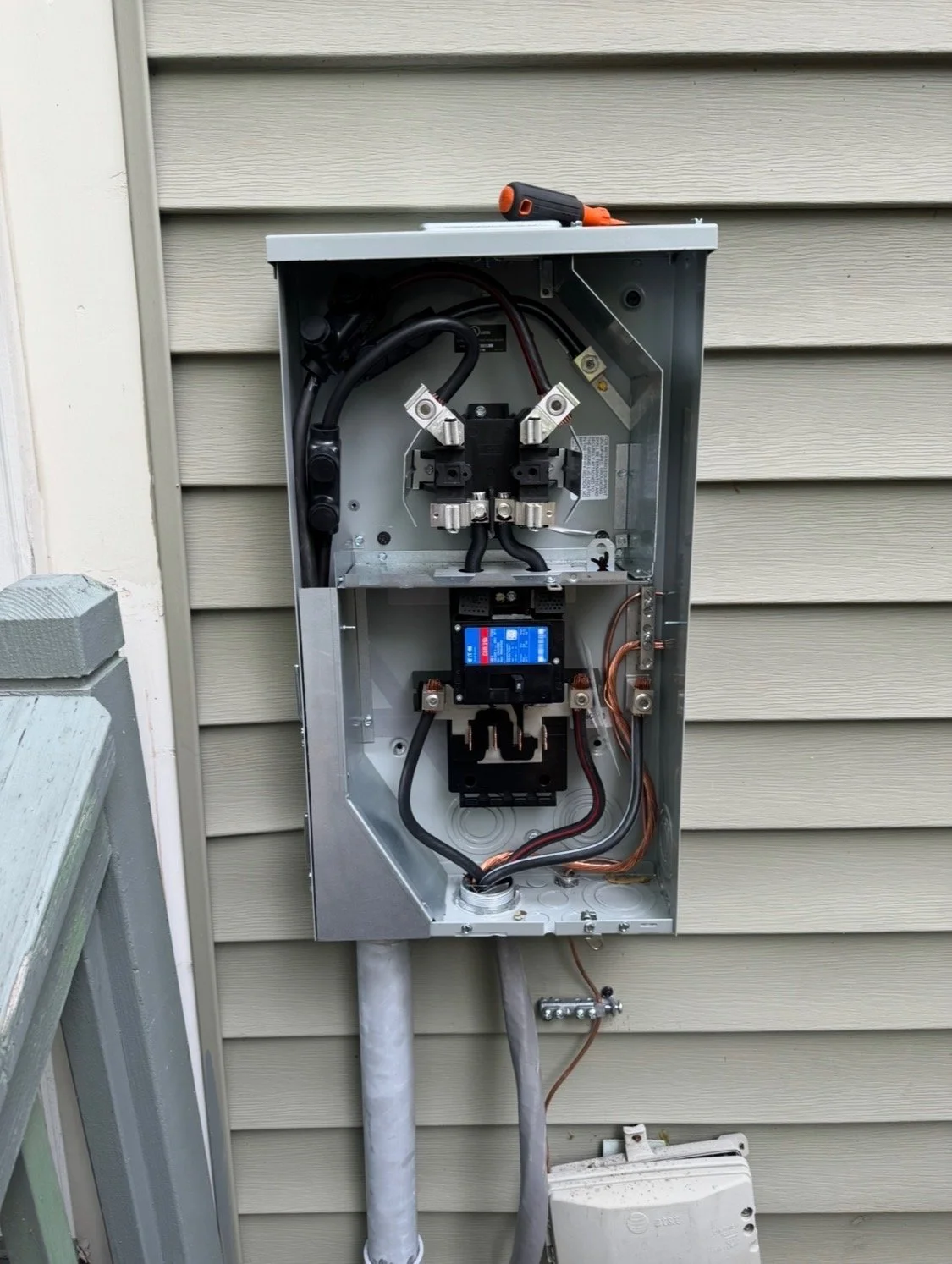Open electrical panel with wires, circuit breakers, and tools outside on beige siding wall.