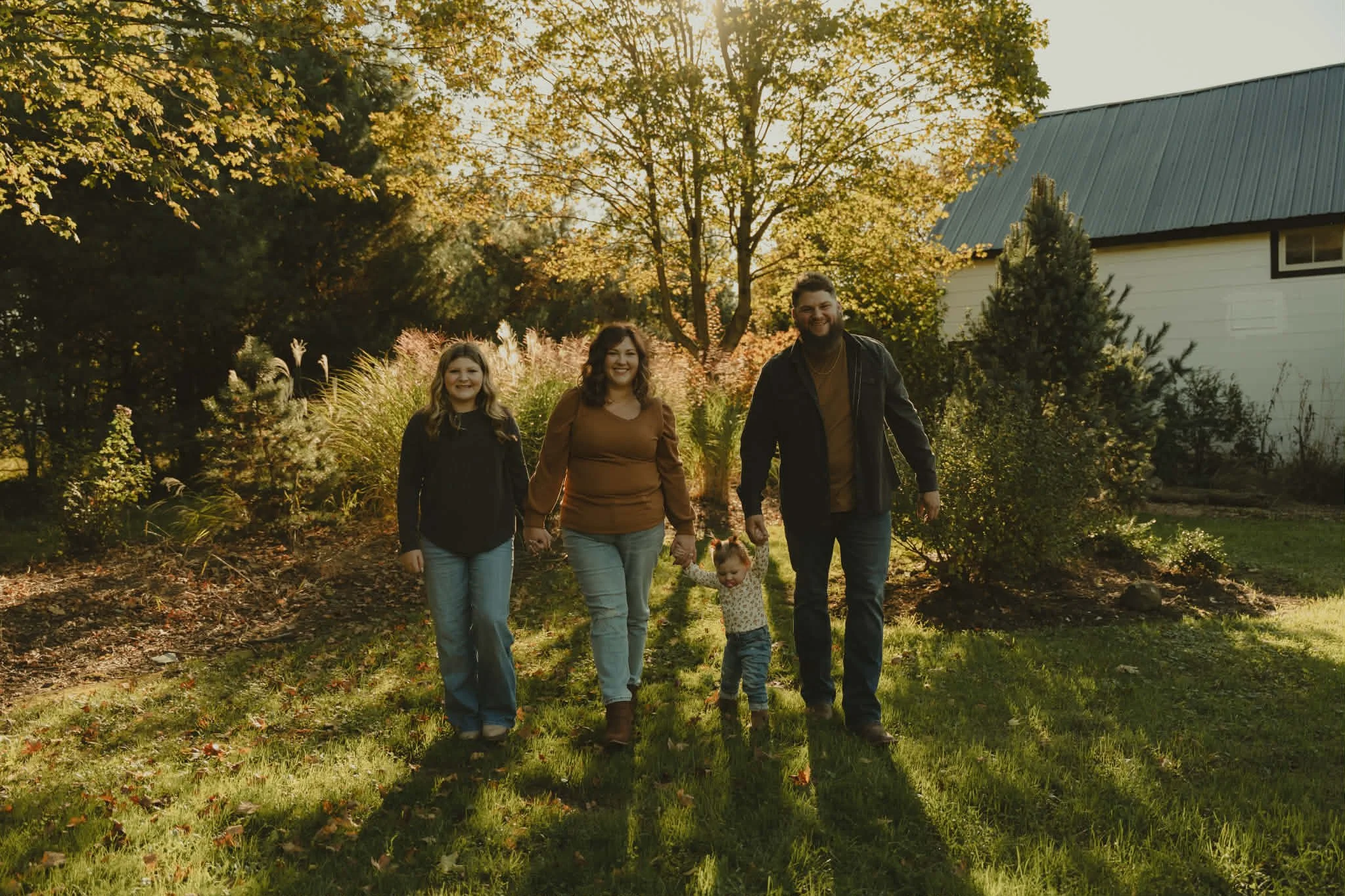 A family walk in a park during autumn, with trees and a house in the background, and sunlight filtering through the leaves.