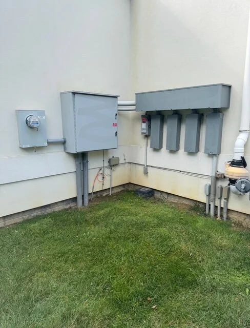 Electrical meters, panels, and conduit outside on the exterior wall of a building with grass in foreground.