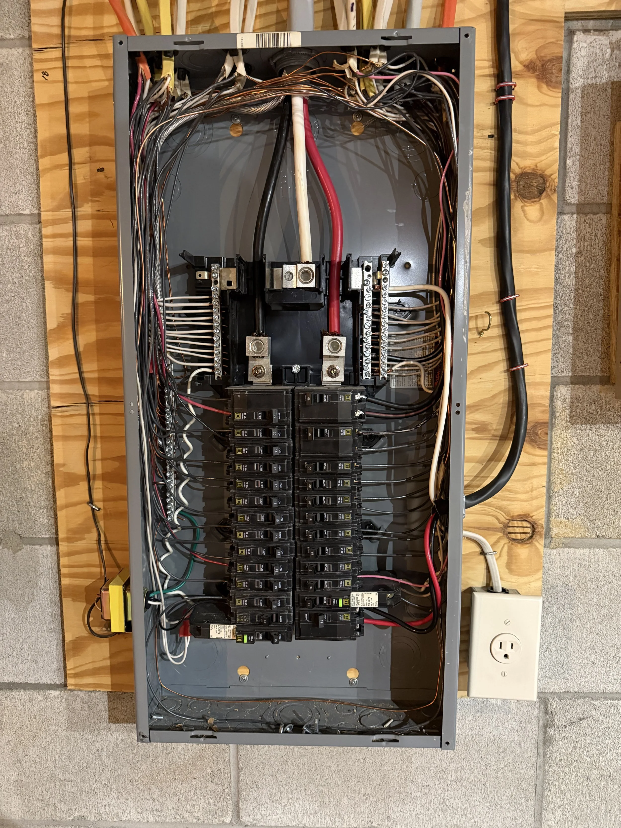 An electrical breaker panel with multiple circuit breakers and wiring mounted inside a metal box, placed on a wooden surface next to a wall with a power outlet.