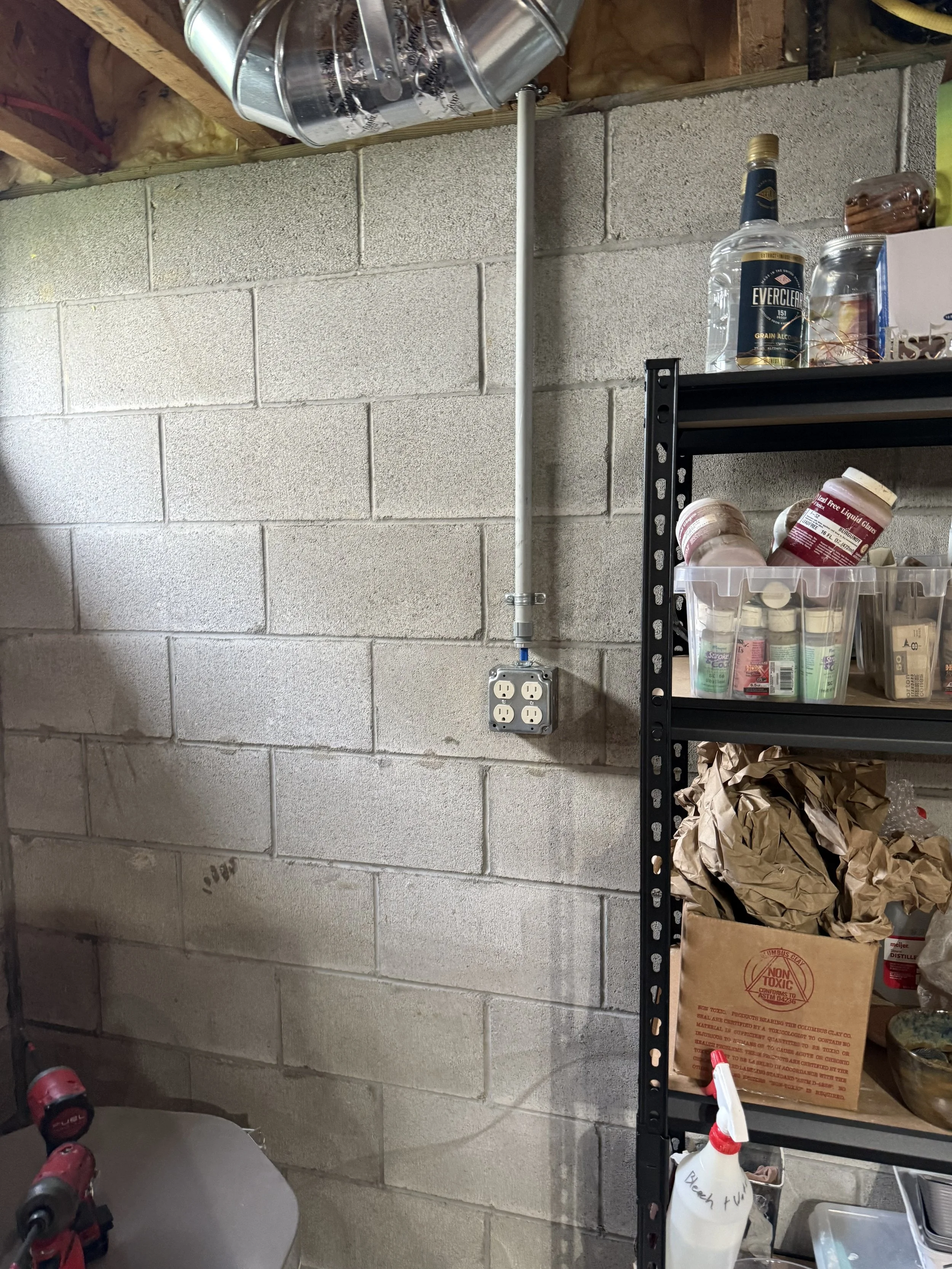 A concrete block wall with an electrical outlet and a pipe running vertically, a black metal shelving unit holding various items including bottles, jars, and paper bags, and a shelf mounted near the ceiling with metal cans.