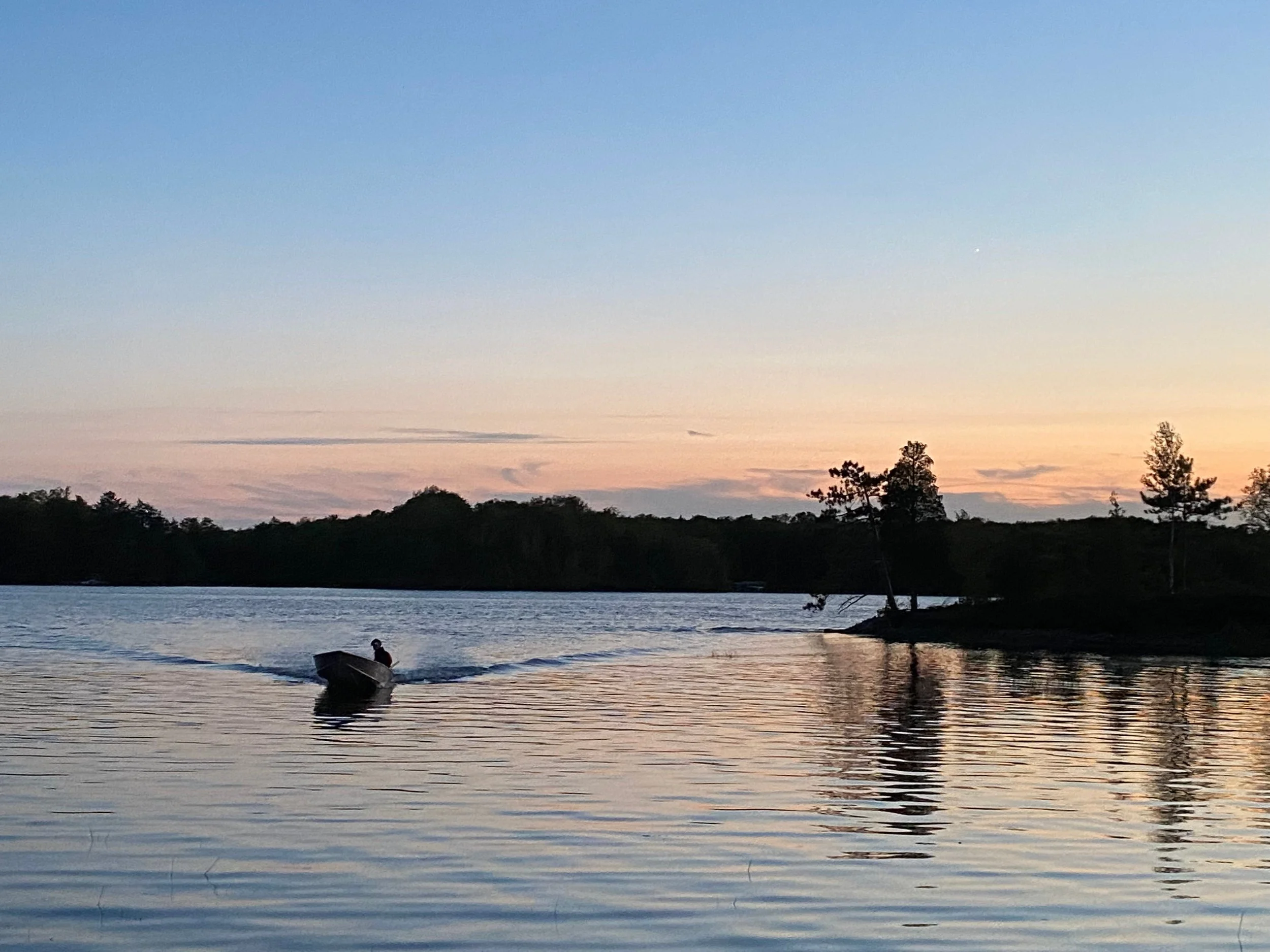 A person in a boat on a river during sunset or sunrise, with trees along the riverbank and a colorful sky.