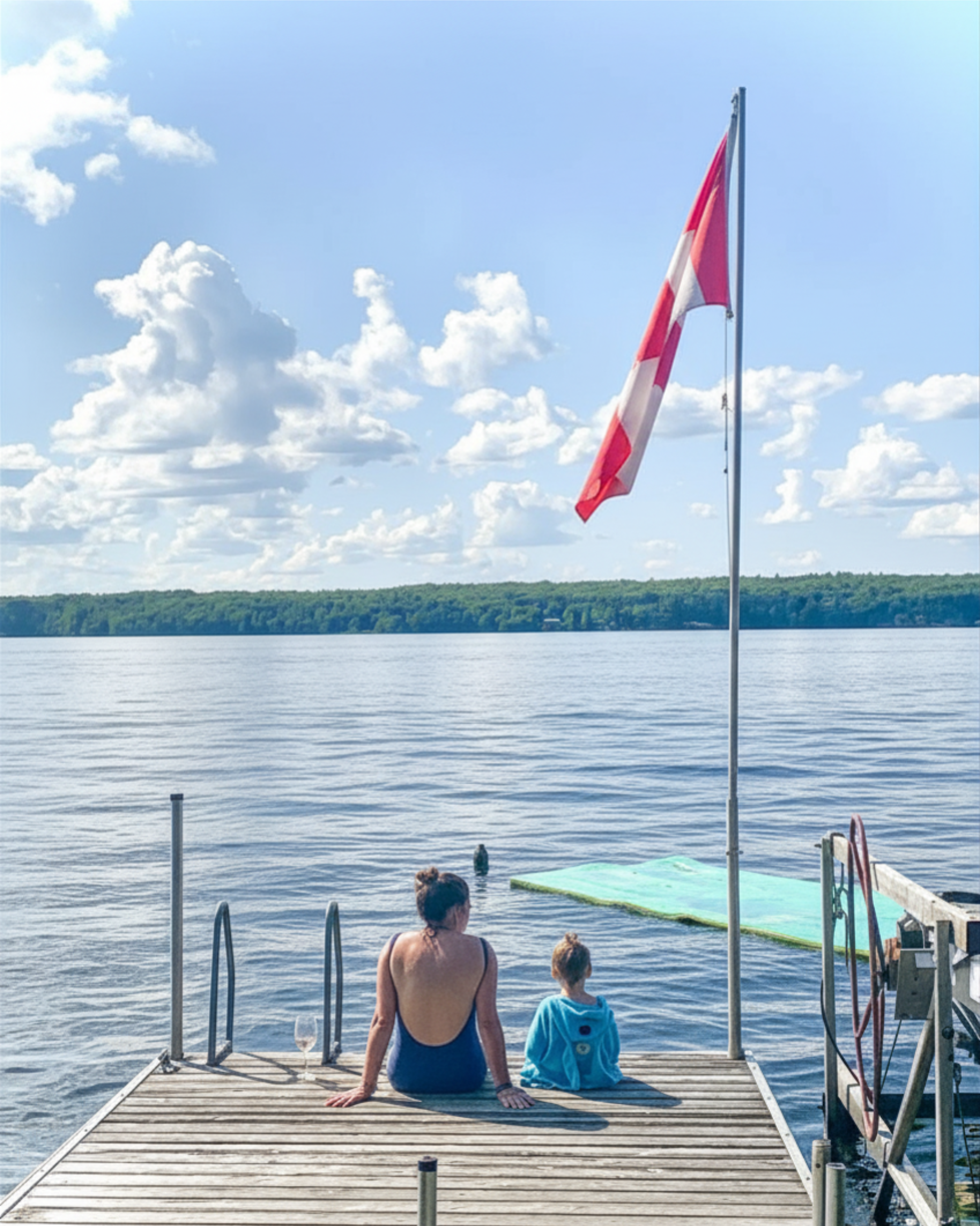 Two people, an adult woman and a young girl, sitting on a wooden dock by a body of water, looking at the water. An American flag is flying on a pole nearby, and the sky is partly cloudy with a green treeline in the background.