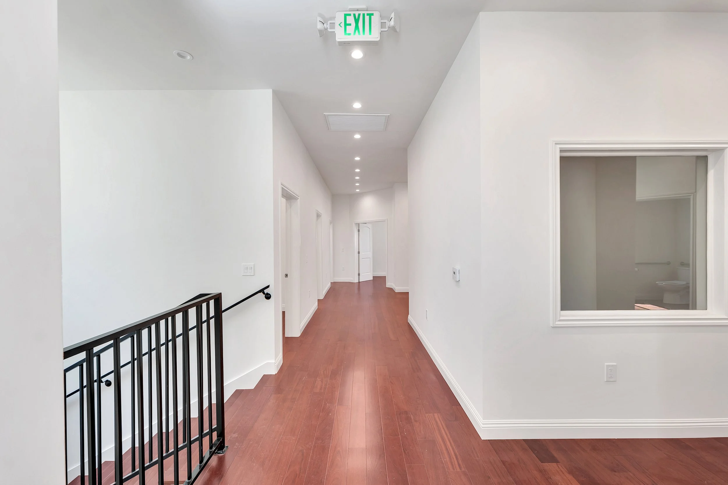 Empty hallway with white walls, wooden floors, and ceiling lights. A black staircase railing is visible on the left, with an illuminated green exit sign hanging on the ceiling. Several doors are visible along the corridor.