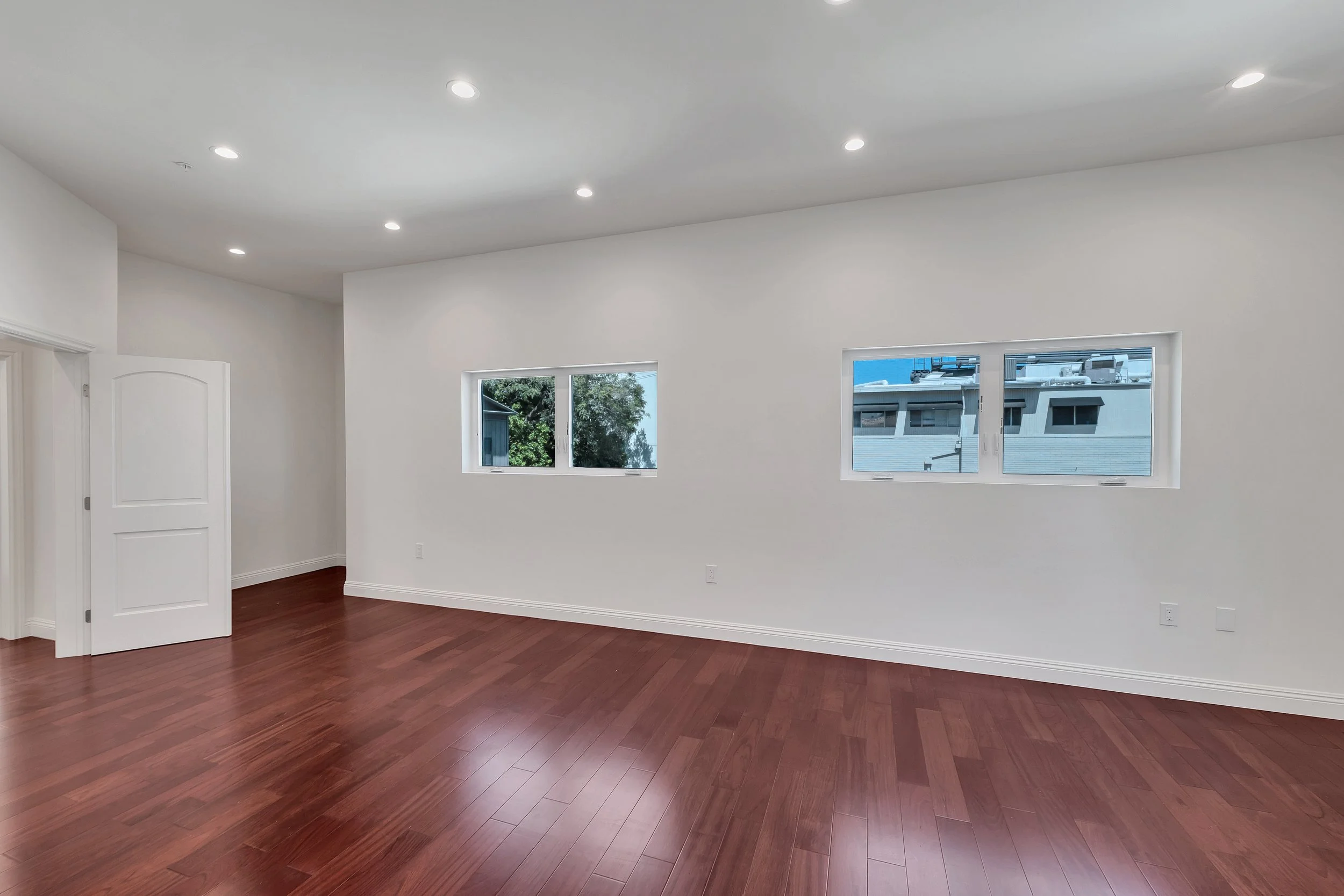 Empty room with white walls, hardwood flooring, and two high windows with trees visible outside.