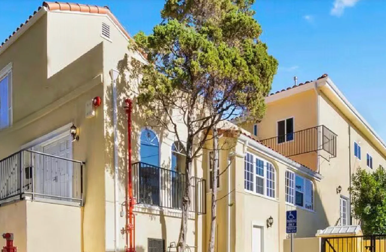 A multi-story yellow residential building with balconies and a tree in front, under a clear blue sky.