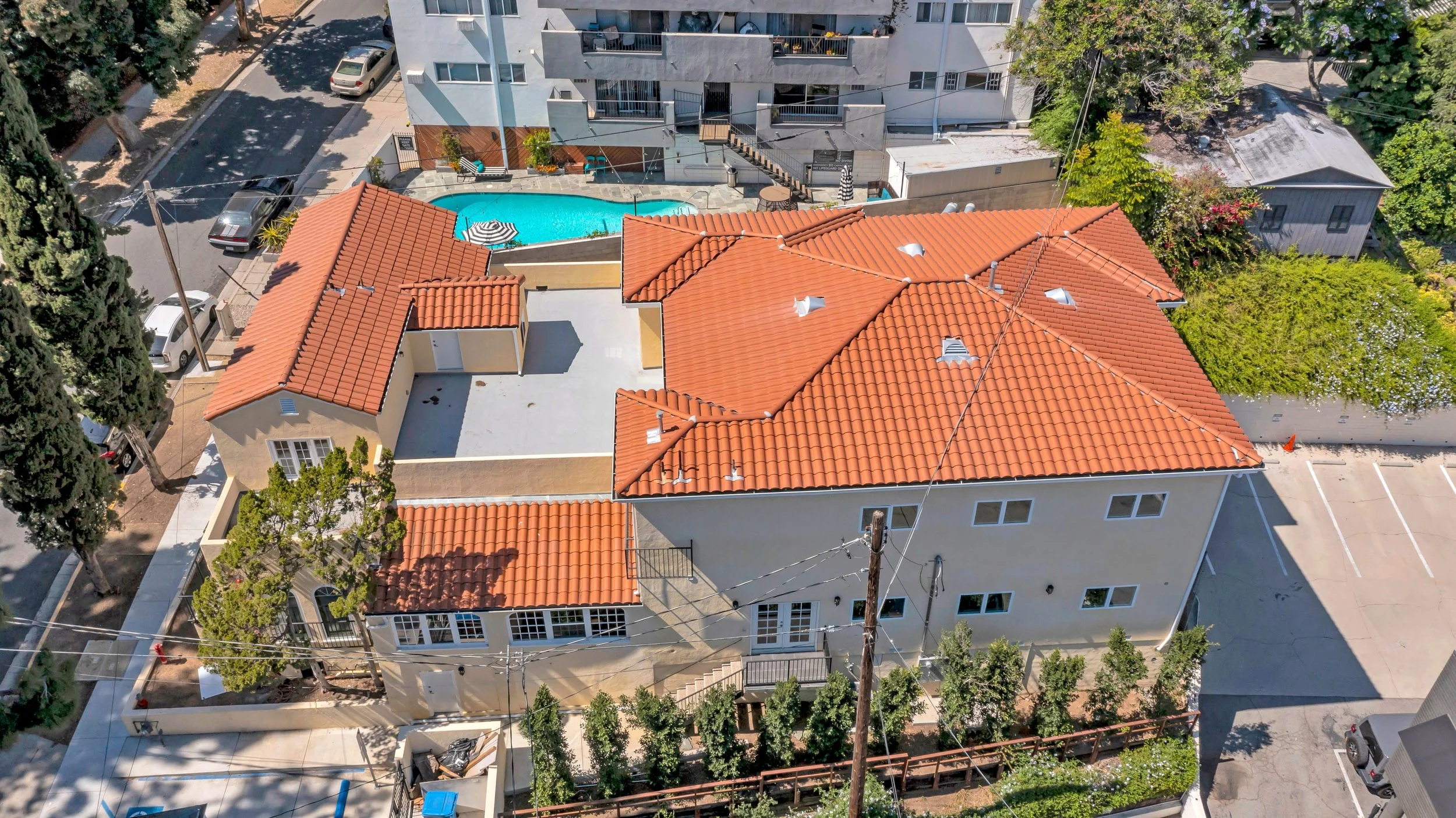 Aerial view of a building with red tile roof, swimming pool, patio umbrellas, surrounding trees, parked cars and nearby streets.