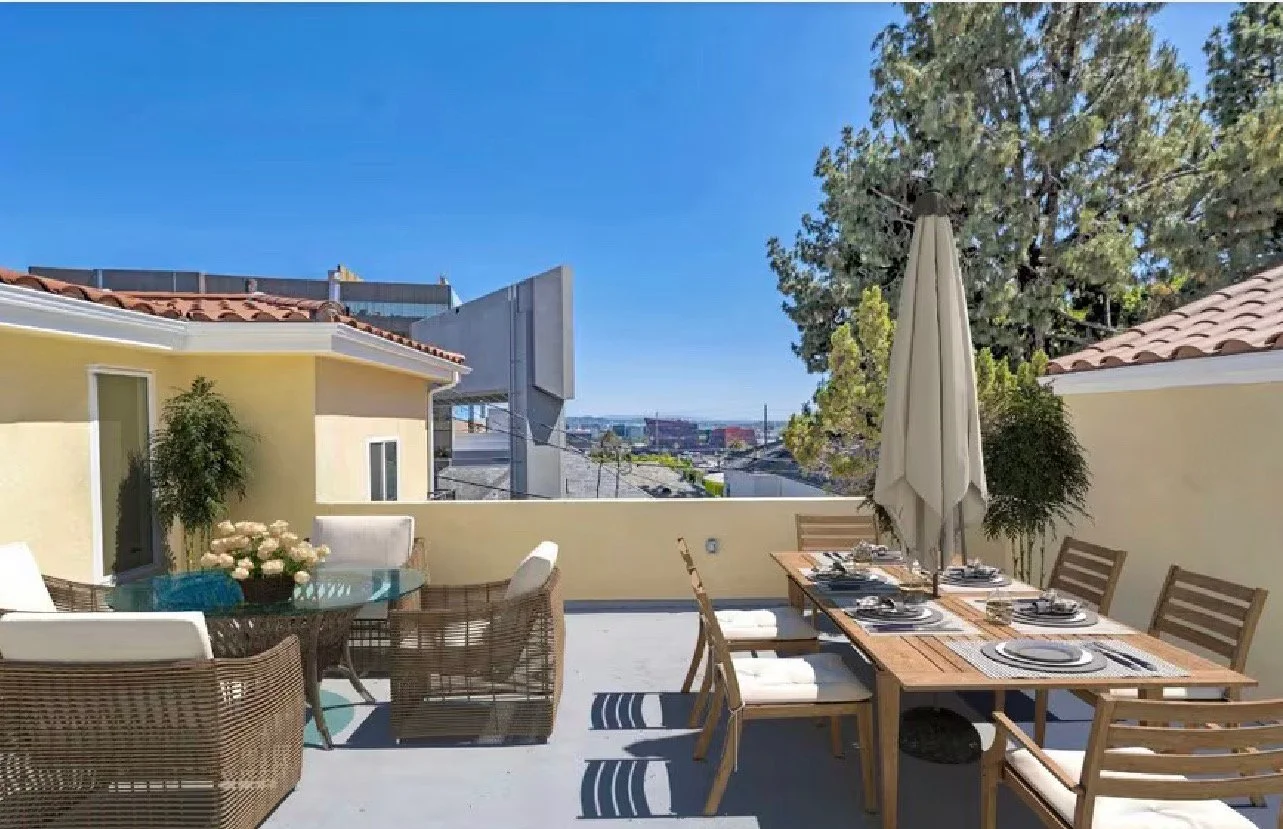 A rooftop patio with a dining table set for a meal, surrounded by chairs, umbrellas, and potted plants, with neighboring buildings and a clear blue sky in the background.