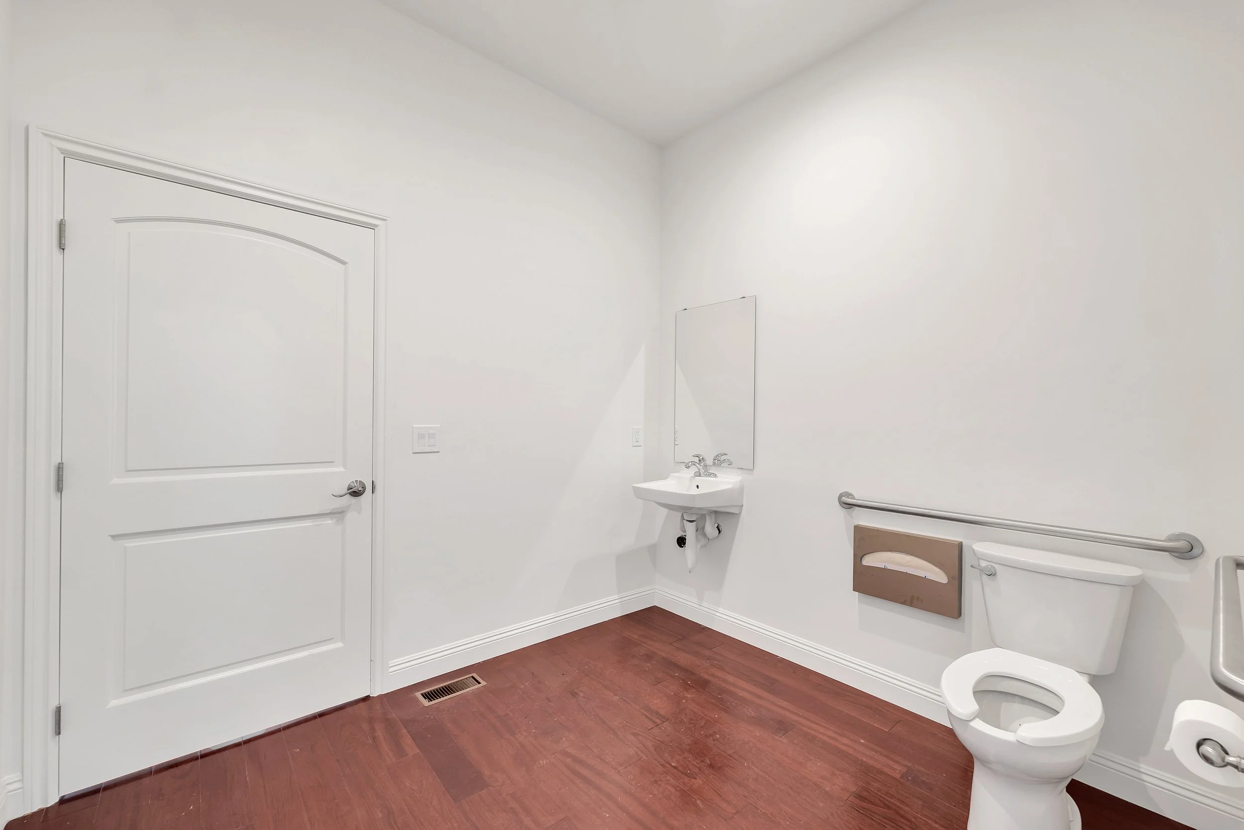 A clean, white bathroom with a closed door, a small wall-mounted sink, a large mirror, a toilet, an emergency call button, a tissue box, a handrail, and a floor vent against warm brown wood flooring.