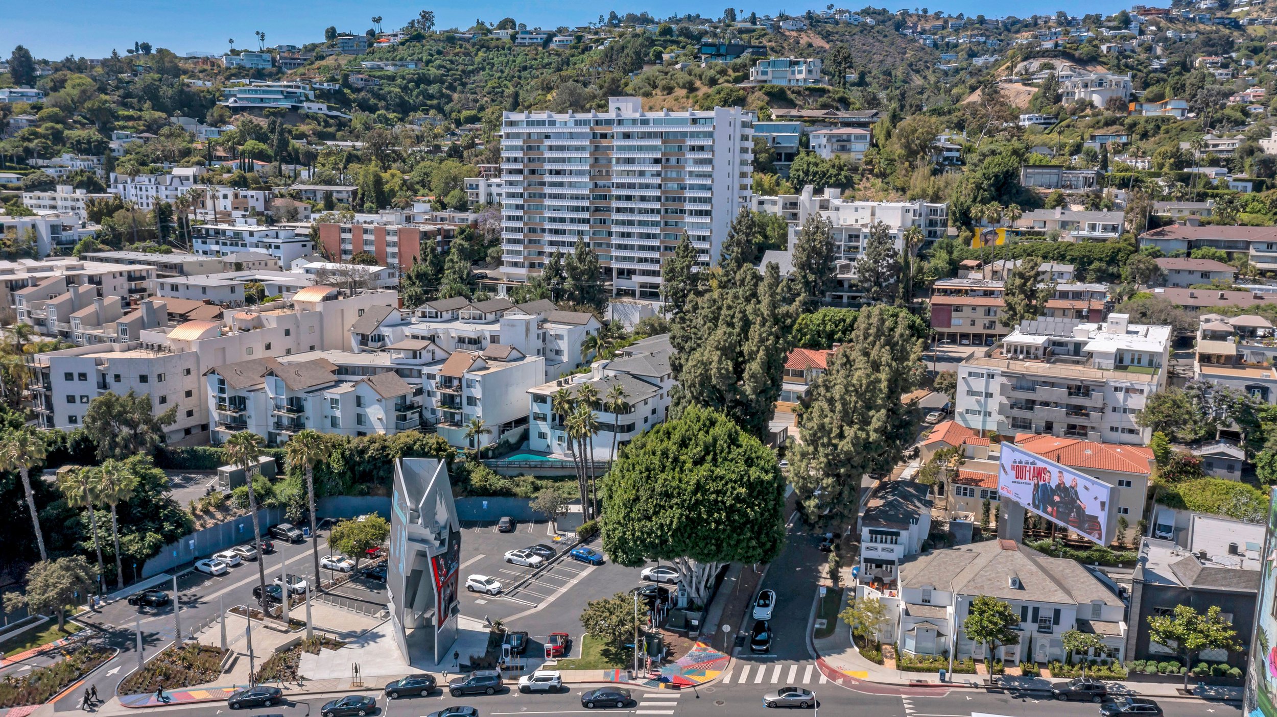 Aerial view of a cityscape featuring a mix of high-rise apartments, mid-rise buildings, and houses on a hillside, with parking lots and streets at street level.