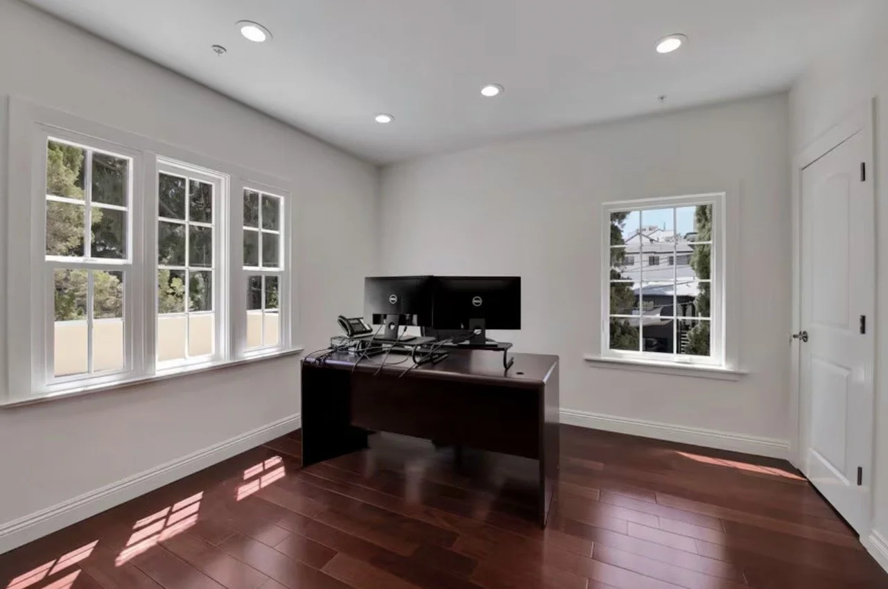 Empty office with white walls, wooden floor, a dark brown desk, and two computer monitors. There are three windows on the left and one window on the right, letting in natural light.