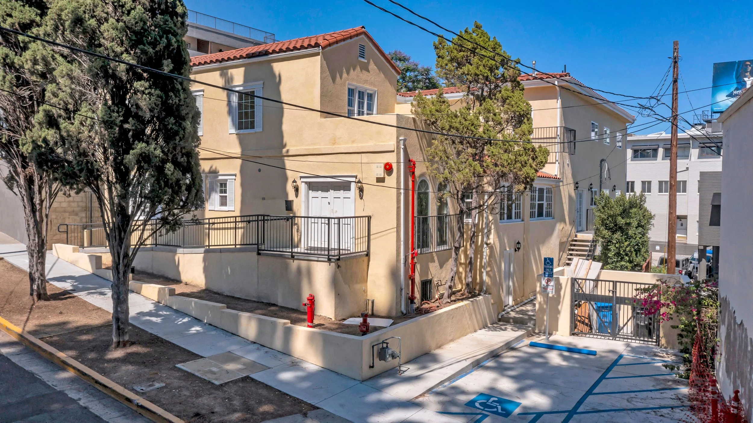 A beige two-story house with white window frames, small balconies, and red tile roof, surrounded by trees on a sunny day with a clear blue sky. There is a parking space marked for handicapped in front of the house.