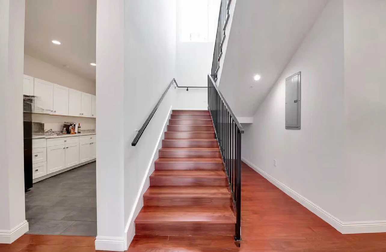Interior view of a staircase with wooden steps, black metal railings, and white walls, next to an open kitchen with white cabinets and dark floor tiles.
