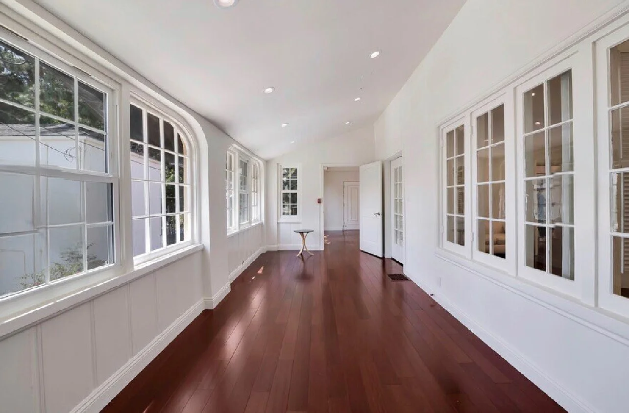 Empty sunroom with white walls, multiple large windows, and hardwood floors, with a small round table near the back.